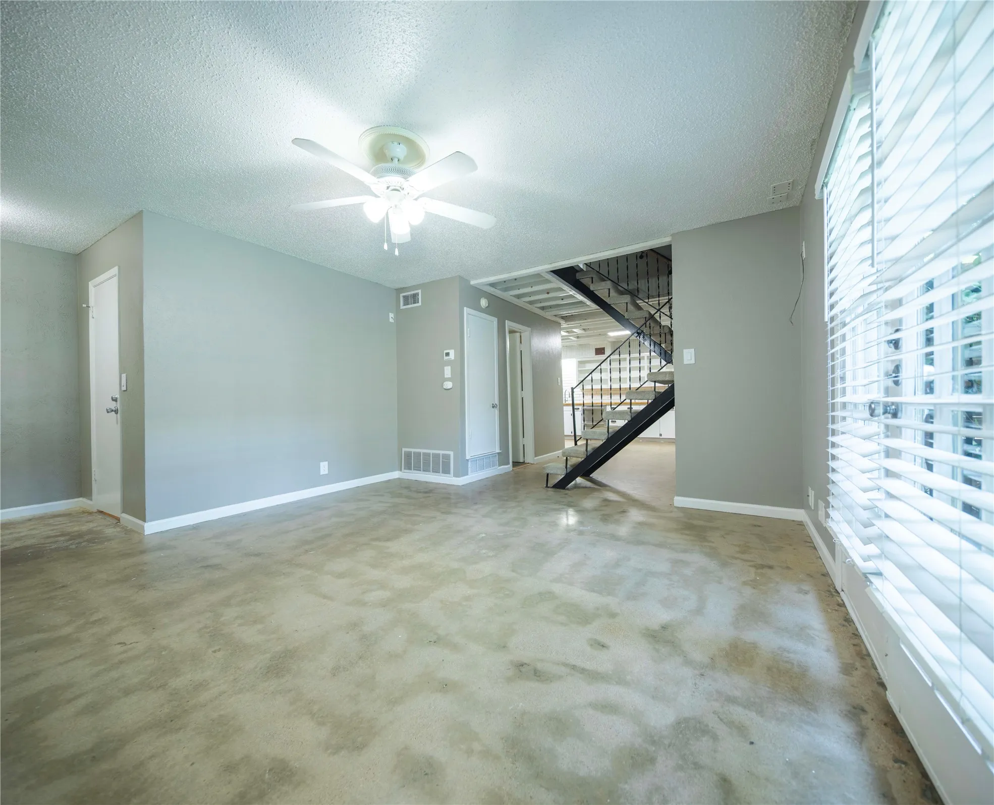 Spacious room featuring light gray wall paint, a ceiling fan with integrated lighting, window blinds for natural light control, and an open staircase with wrought iron railing