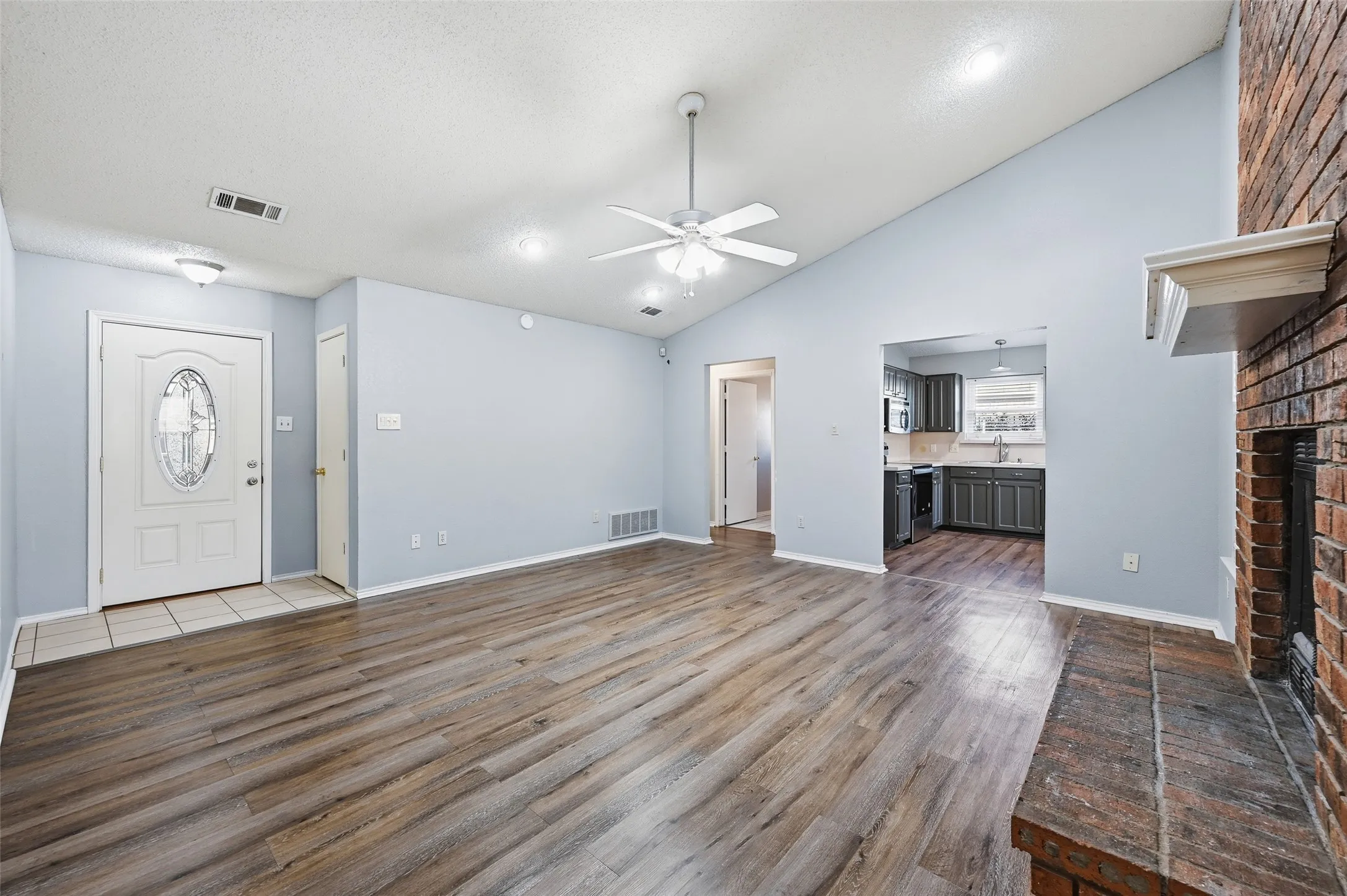 Vaulted ceiling with a ceiling fan, wood-finish flooring, brick fireplace with a mantel, and a kitchen with dark cabinetry