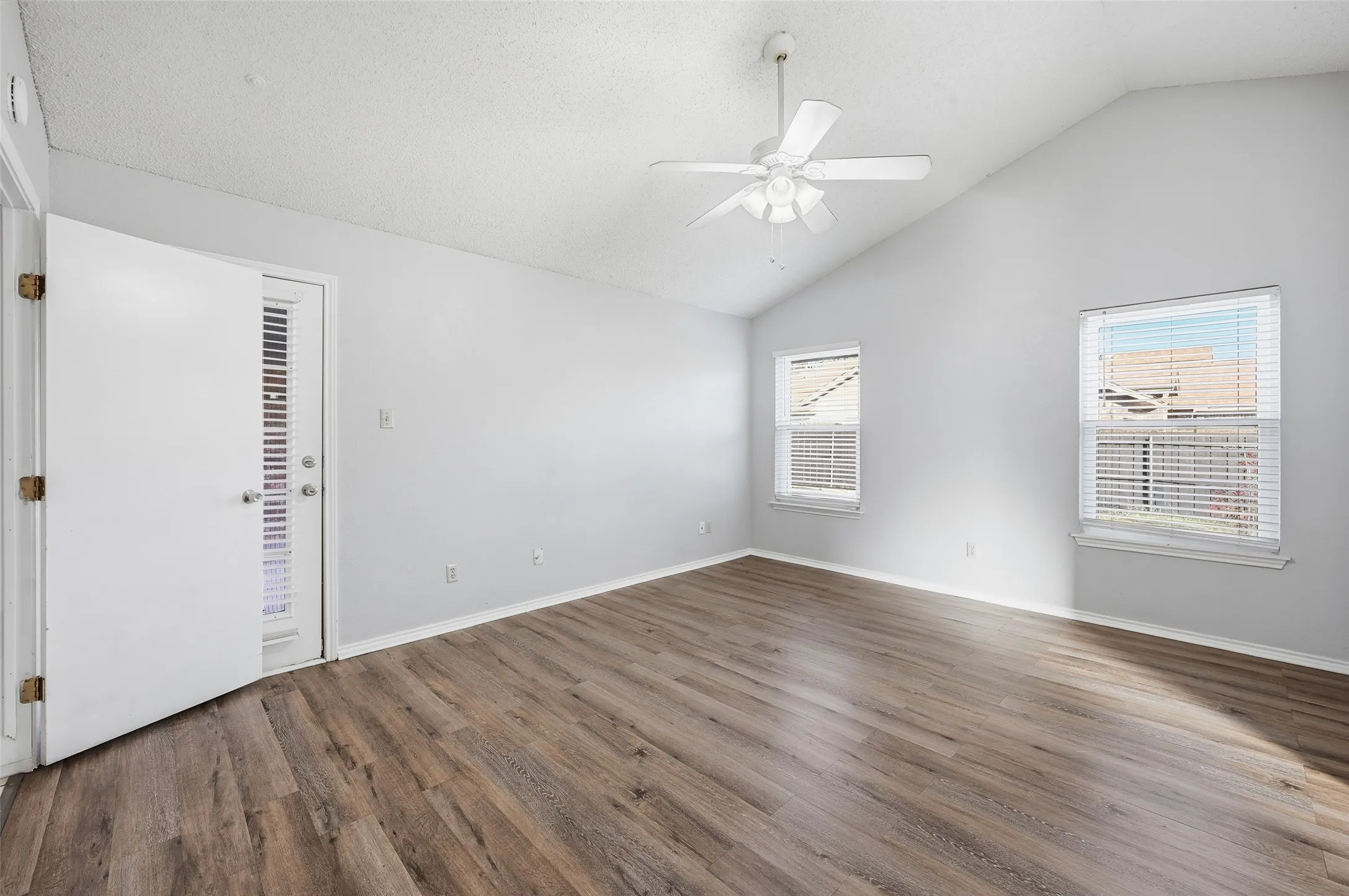 Vaulted ceiling with a ceiling fan, wood-finish flooring, two windows with blinds, and a door with an integrated blind