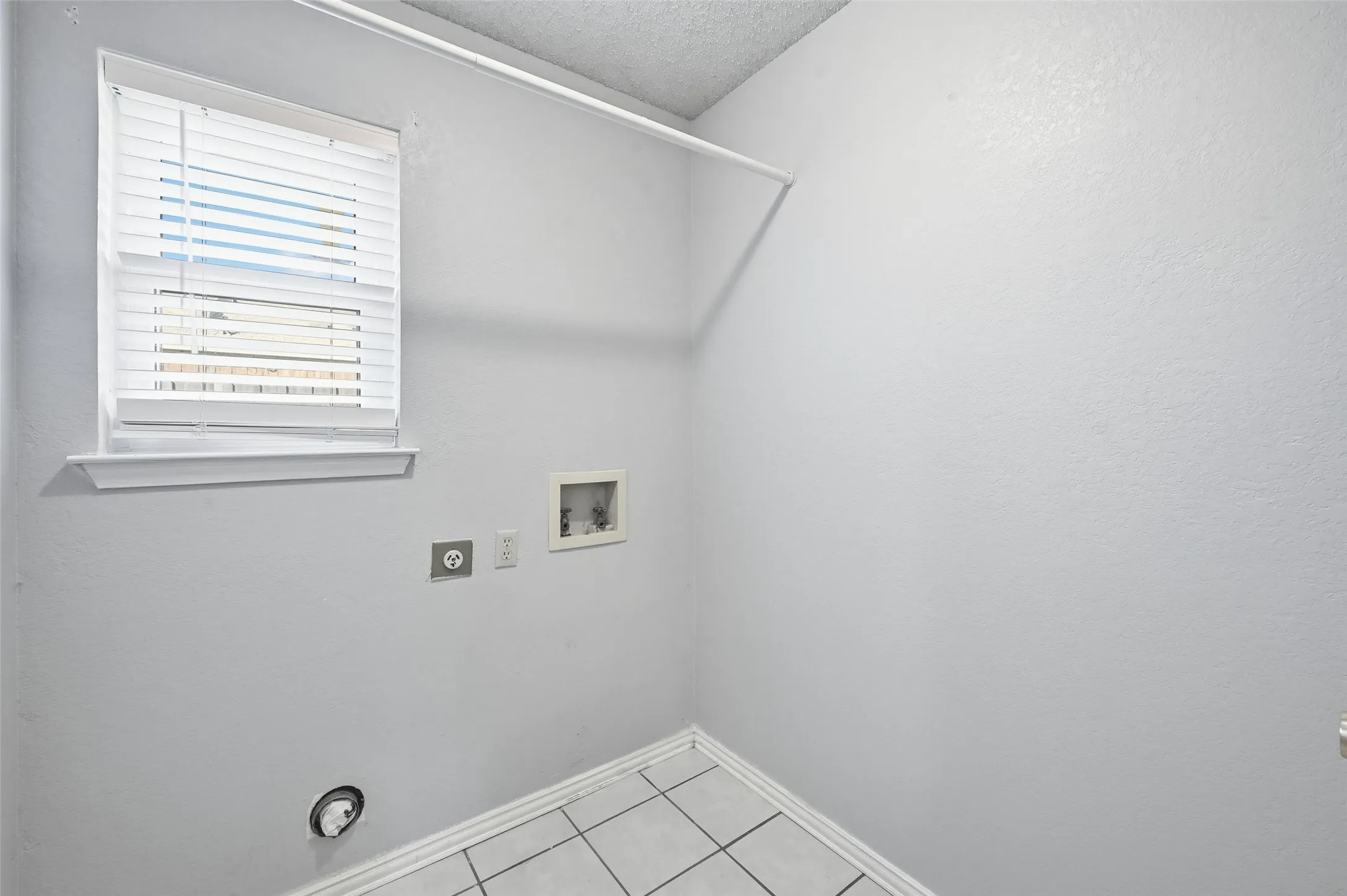 White wall paint, window with blinds, tiled flooring, wall-mounted laundry hookups, and a clothes hanging rod
