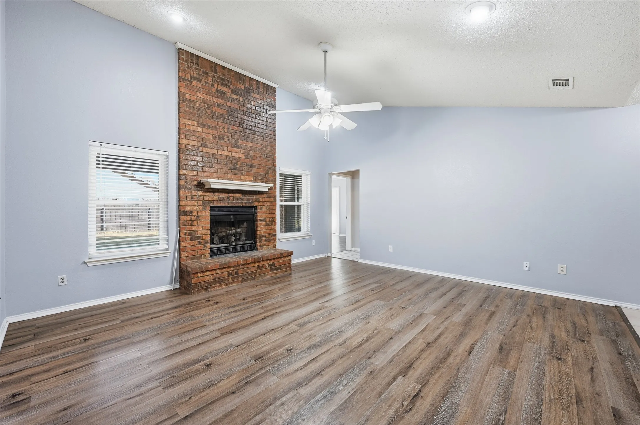 Spacious room featuring vaulted ceilings, wood-finish flooring, a brick fireplace with a mantel, and dual windows