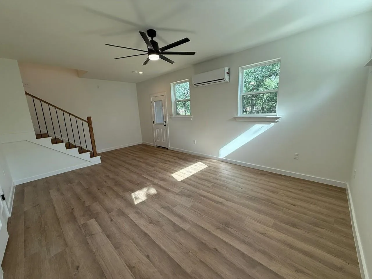 Spacious room featuring wood-finish flooring, white walls, multiple windows, and a modern ceiling fan with integrated lighting