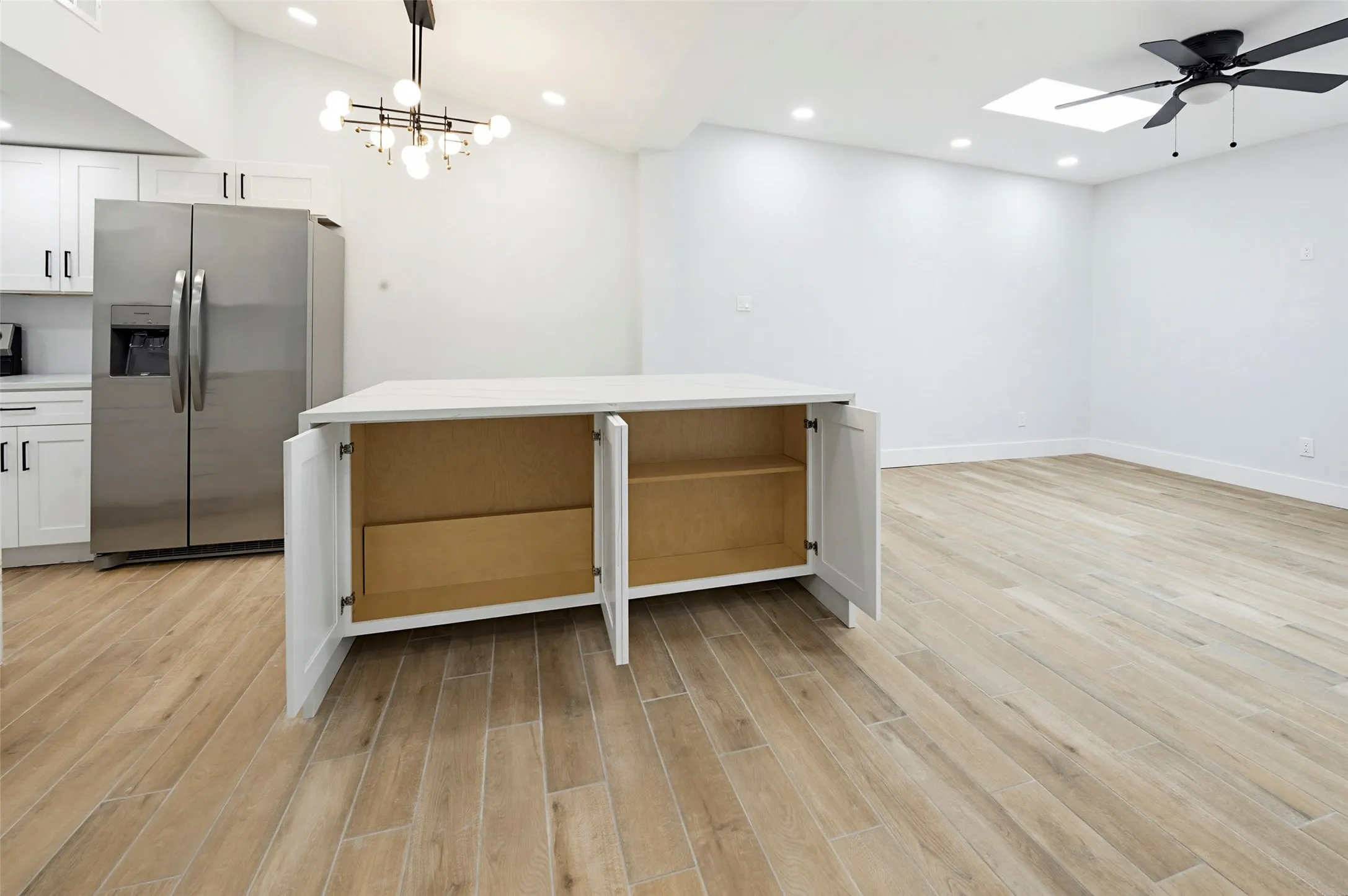 Open-concept room featuring wood-finish flooring, a kitchen island with storage, stainless steel refrigerator, contemporary chandelier, and a ceiling fan with a skylight