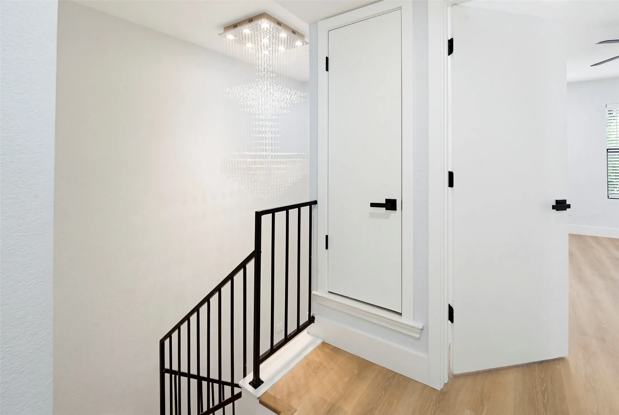 Contemporary chandelier illuminating the stairwell, black metal stair railing, light wood-finish flooring, and white interior doors with matte black hardware