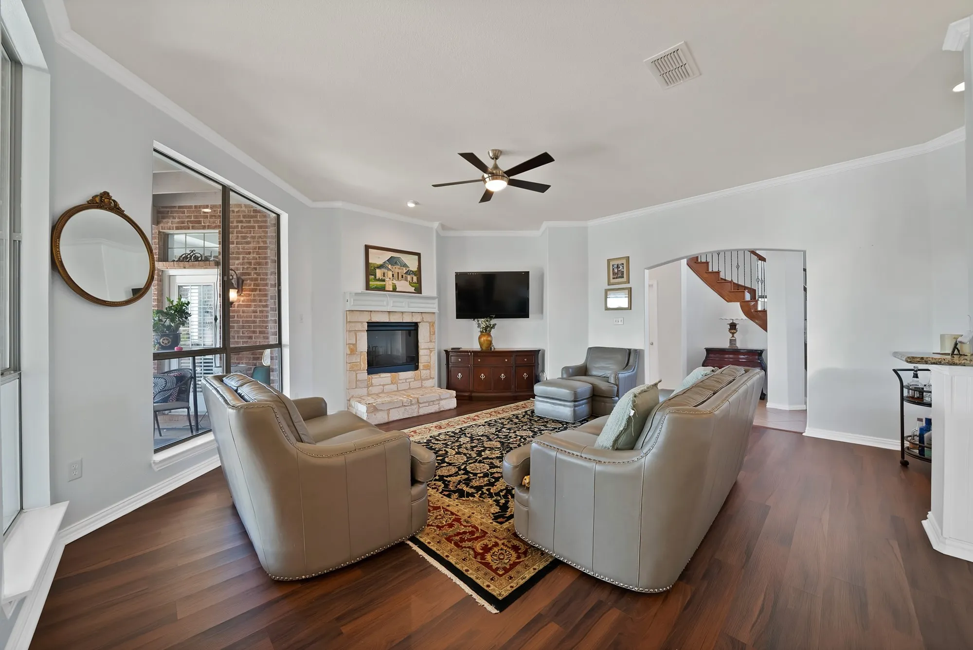 Family room — Stone surround fireplace with raised hearth, dark hardwood floors, crown molding, ceiling fan, and oversized sliding glass doors with direct access to covered back patio.