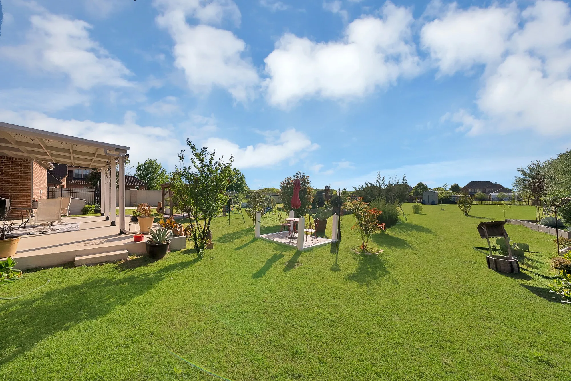 Rear yard, aerial view — Expansive rear yard with solar panel array on roofline, covered pergola structure, raised garden beds, mature trees and ornamental plantings, iron gate access, and upper-level covered balcony overlooking the property.