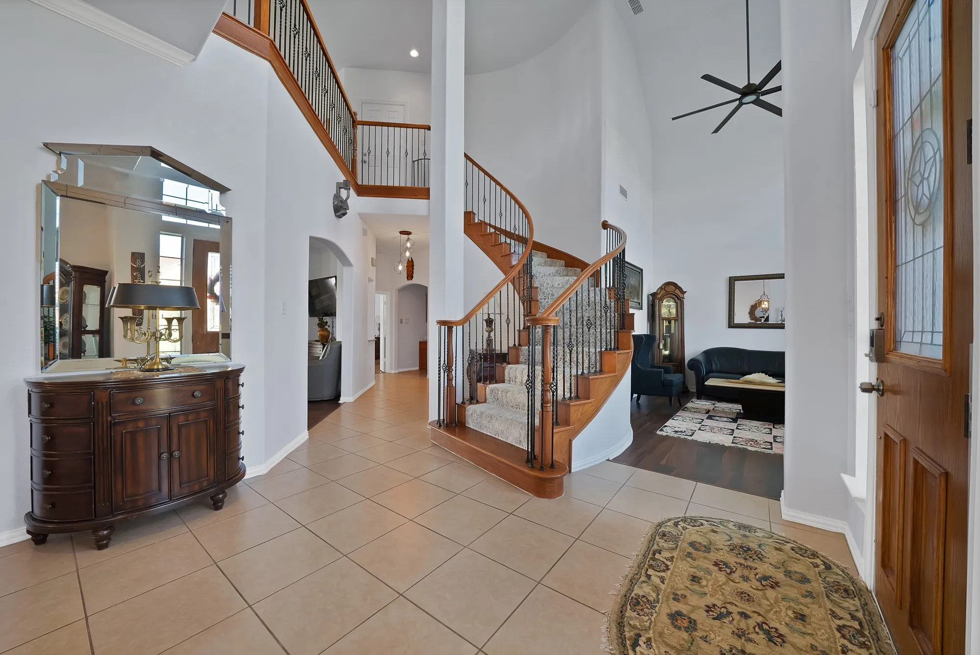 Entry foyer — Two-story rotunda entry with curved hardwood staircase, decorative wrought iron balusters, large-format tile flooring, leaded glass sidelight panels flanking the front door, and open sightlines to both the formal living area and the main corridor.