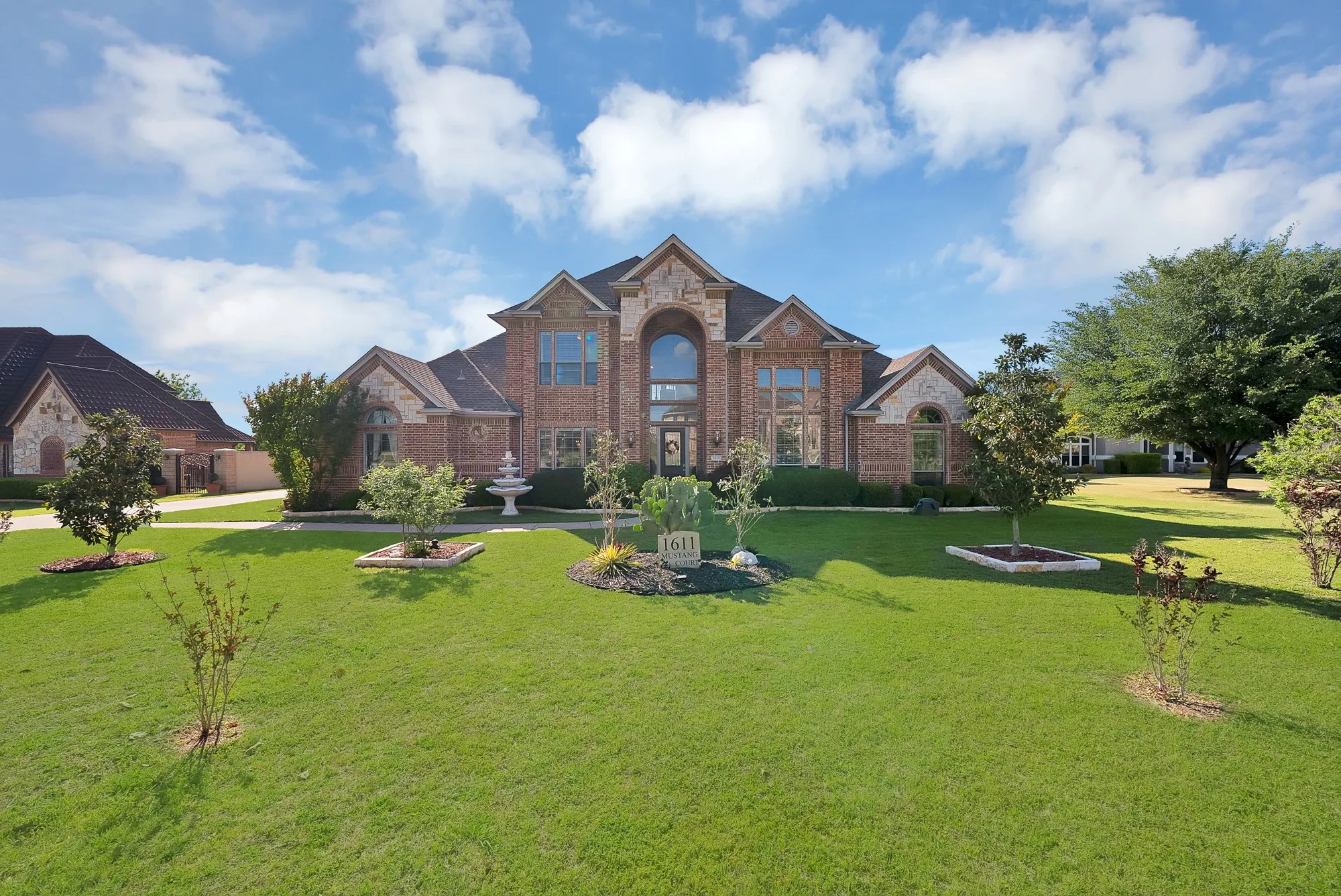 Front exterior, drone view — Two-story brick and cut stone facade with arched entryway, two-story window wall, multi-gabled roofline, tiered fountain centerpiece, and stone-edged landscape beds across a deep front yard. Solar panels visible on rear roofline.