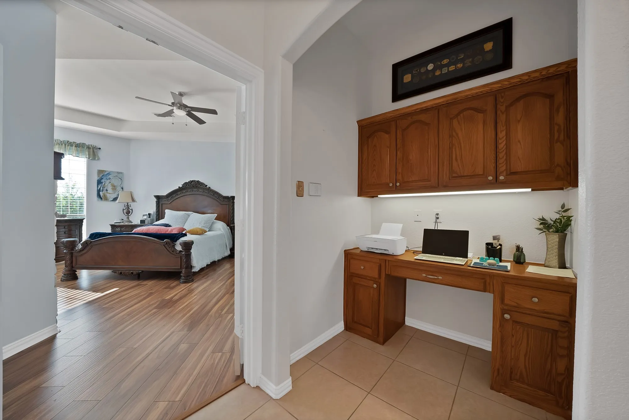 Primary suite sitting area and built-in desk — Built-in oak desk with upper cabinetry, under-cabinet lighting, tile flooring, and arched passthrough to primary bedroom with