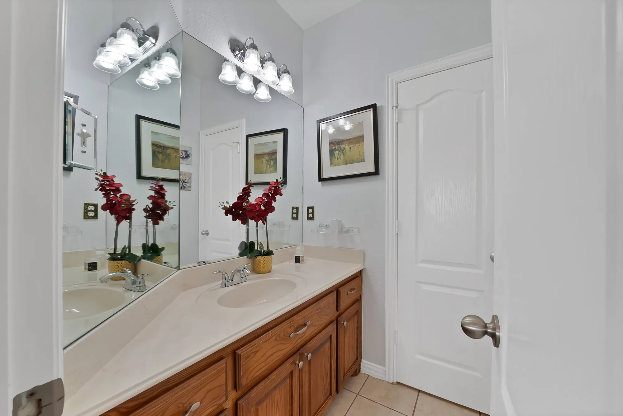 Secondary bathroom, dual vanity — Dual-sink vanity with oak cabinetry and white countertop, full-width mirror, chrome fixtures, multi-bulb vanity bar light, large-format tile flooring, and separate water closet.