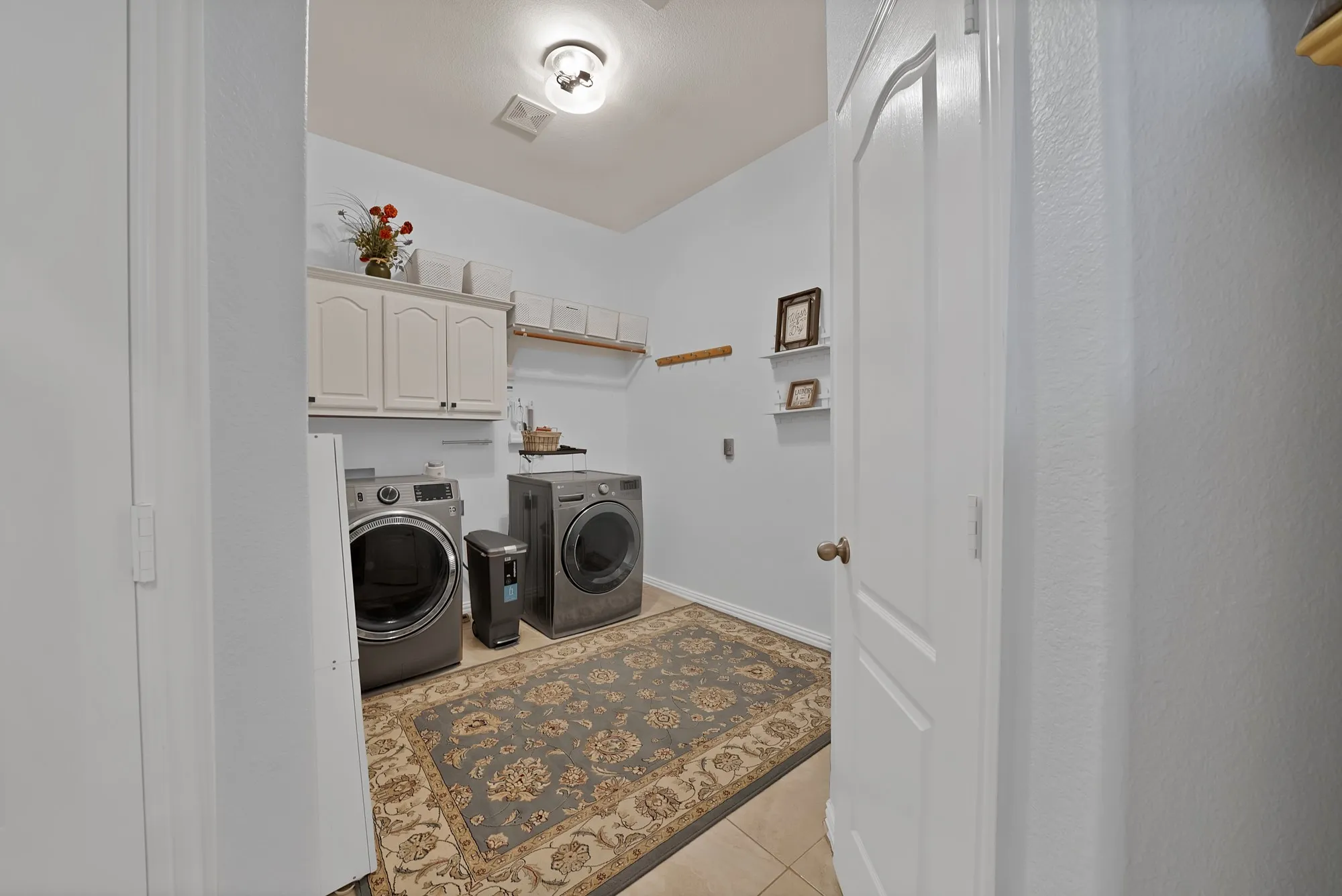 Utility room — Full-size front-load washer and dryer, upper cabinetry with arched panel doors, open wood shelving with hanging rod, and large-format tile flooring.