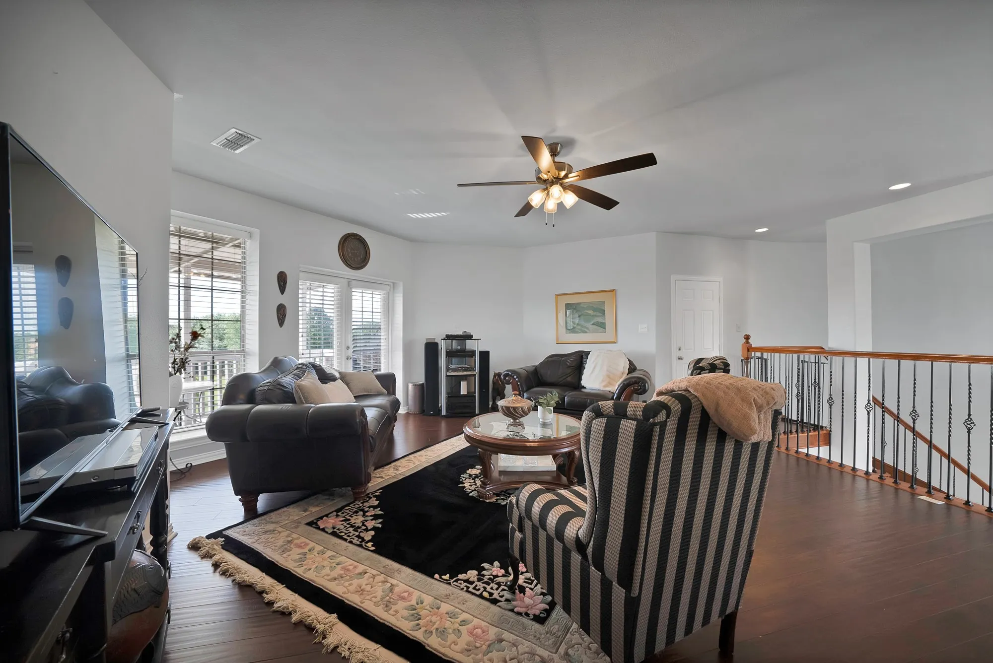 Upper-level living area — Dark hardwood floors, ceiling fan with light kit, French doors to upper balcony, and open sightline to staircase with wrought iron baluster railing. Room functions as a second living or flex space.