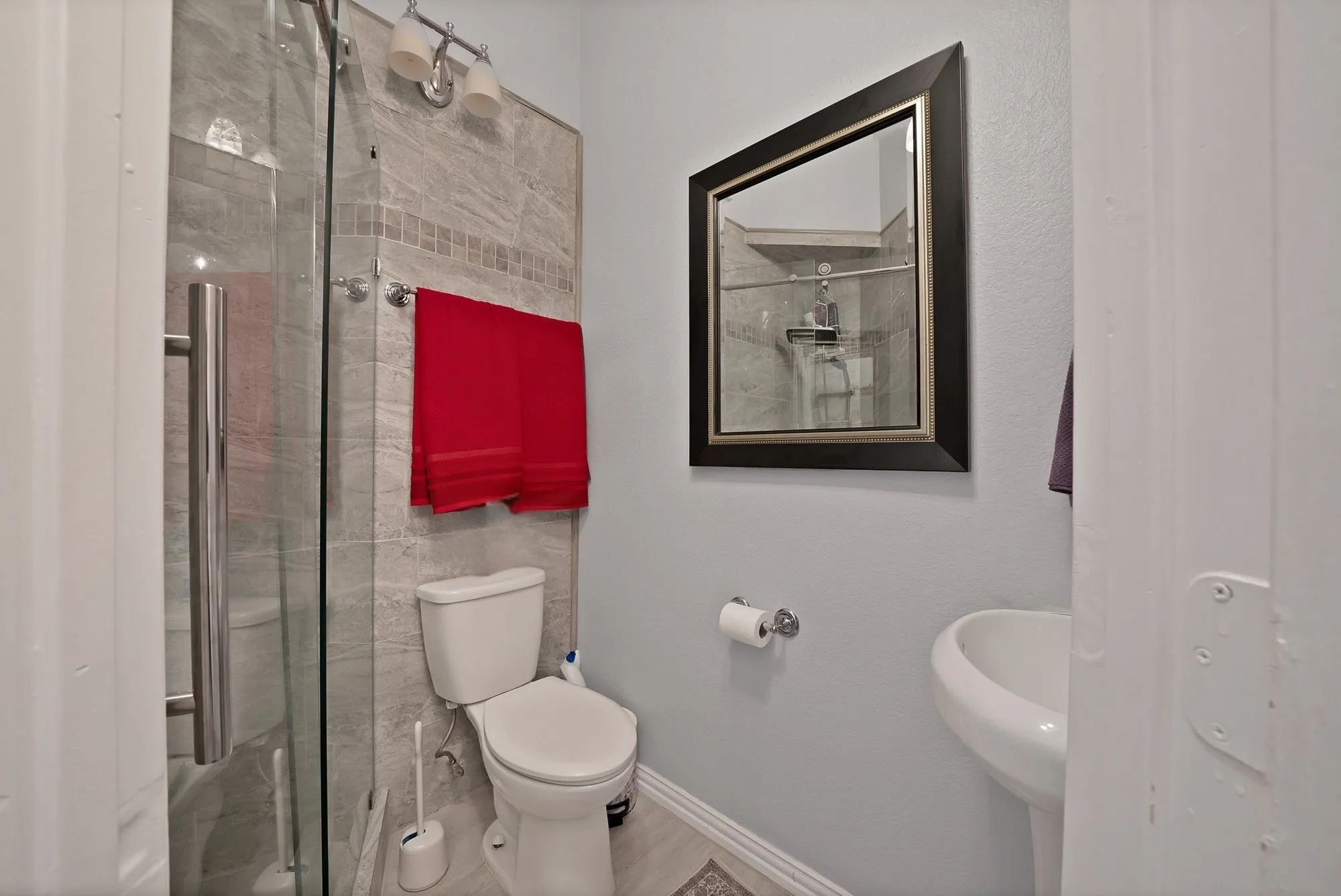 Secondary bathroom, shower only — Frameless glass walk-in shower with floor-to-ceiling gray tile surround and accent mosaic border. Pedestal sink and chrome fixtures visible.