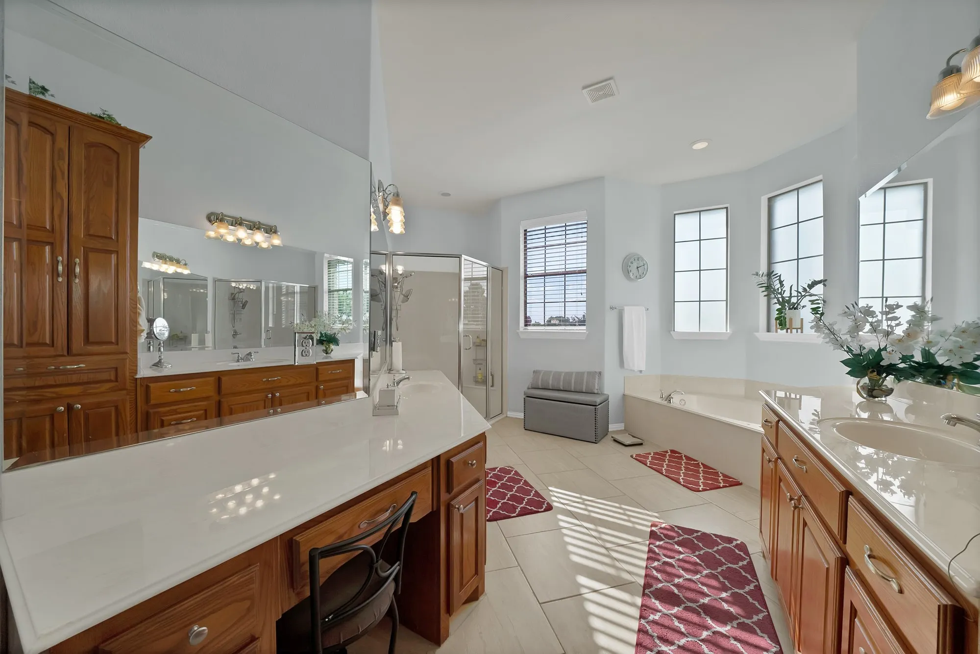 Primary bathroom — Dual oak vanities with white quartz countertops, built-in makeup vanity with knee space, frameless glass walk-in shower, garden tub with tile surround, large-format tile flooring, and triple window bank with plantation blind treatments.
