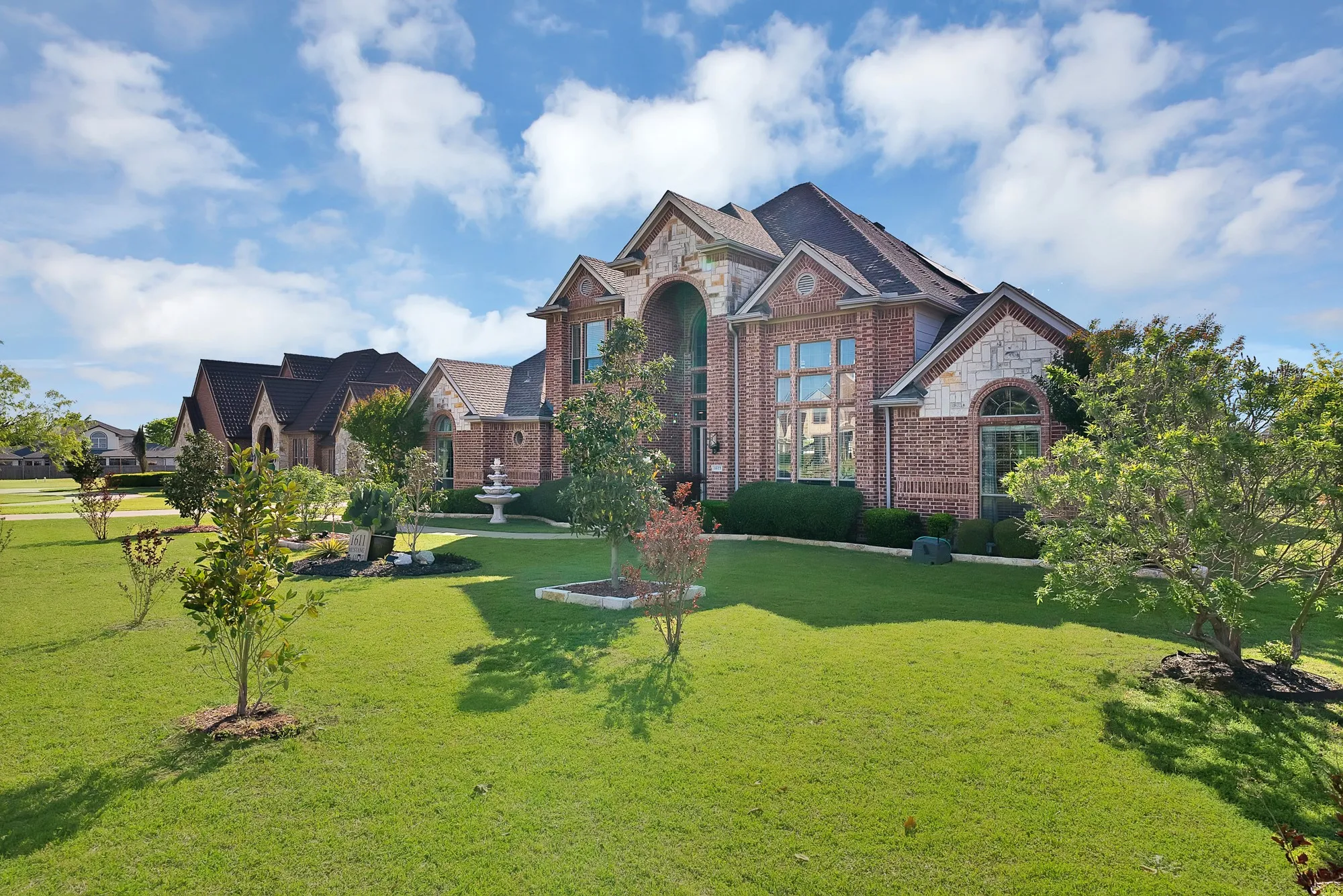 Front exterior, side angle — Full front yard depth visible with circular fountain centerpiece, stone-framed planting beds, mature shade trees, and covered side entry.