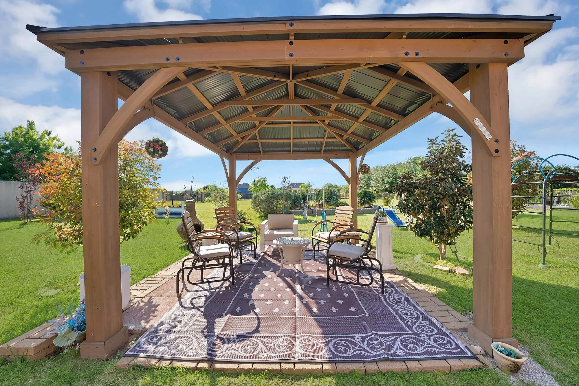 Backyard gazebo — Cedar-frame gazebo with metal hip roof, exposed rafter ceiling, brick-paver base, and fire pit seating area. Set within mature landscaped yard with iron fencing and established ornamental trees visible beyond.