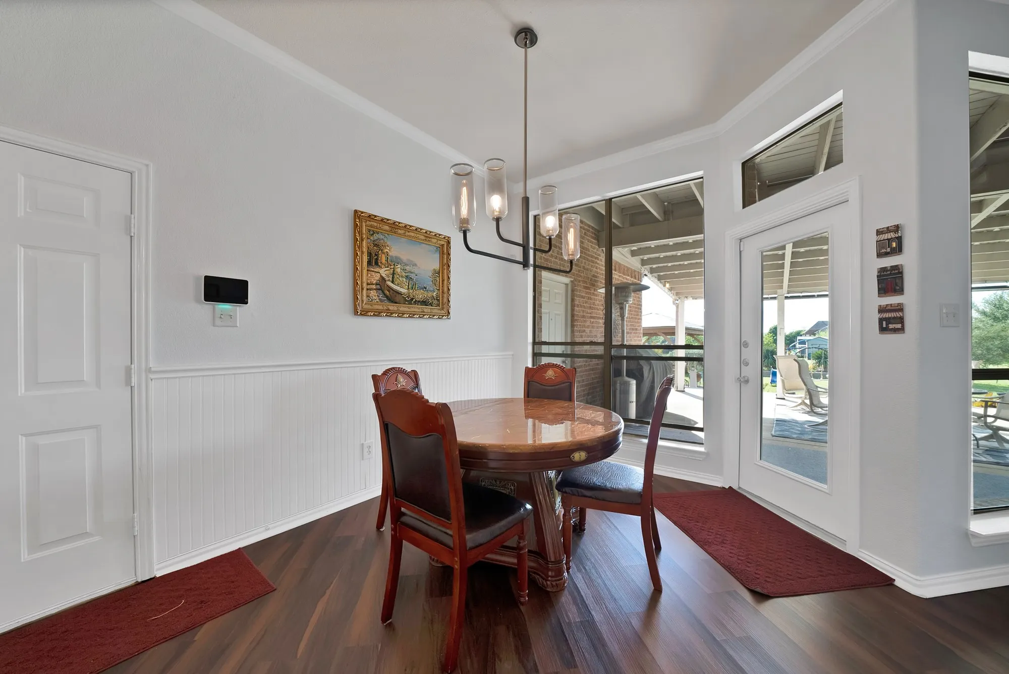 Breakfast nook — Beadboard wainscoting, dark hardwood floors, tray ceiling with crown molding, modern industrial pendant chandelier, smart home panel, and direct door access to covered back patio.