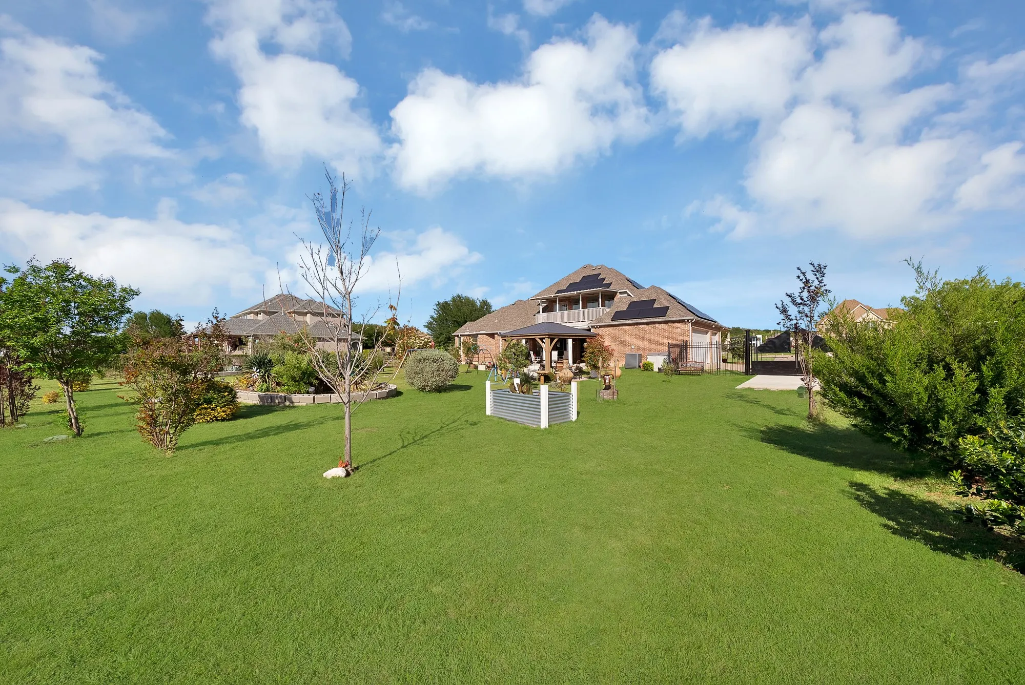 Rear yard, aerial view — Expansive rear yard with solar panel array on roofline, covered pergola structure, raised garden beds, mature trees and ornamental plantings, iron gate access, and upper-level covered balcony overlooking the property.