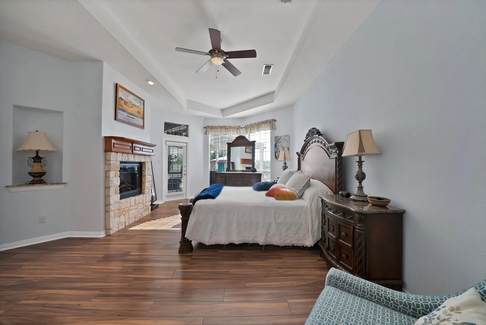Primary bedroom, fireplace view — Tray ceiling, stone surround fireplace with raised hearth and wood mantel, dark hardwood floors, built-in display niche with granite shelf, and direct door access to back patio.