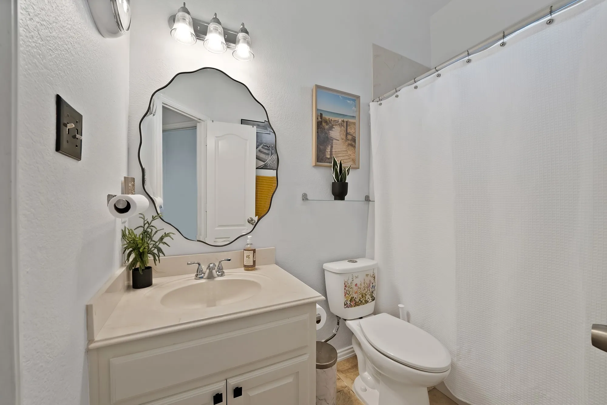 Secondary bathroom — Single-sink vanity with white raised-panel cabinetry, chrome fixtures, decorative mirror, three-bulb vanity bar light, tile flooring, and tub-shower combination.