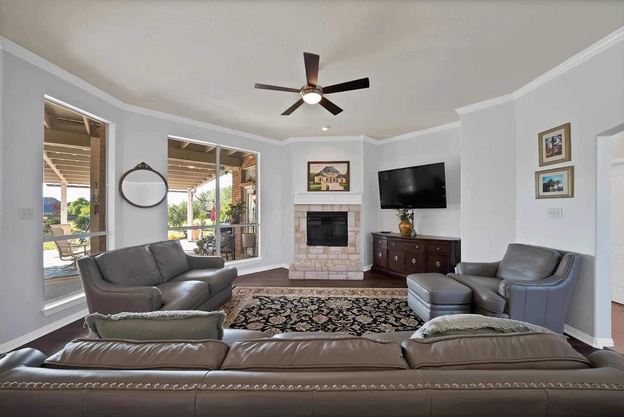 Family room — Stone surround fireplace with raised hearth, dark hardwood floors, crown molding, ceiling fan, and oversized sliding glass doors with direct access to covered back patio.