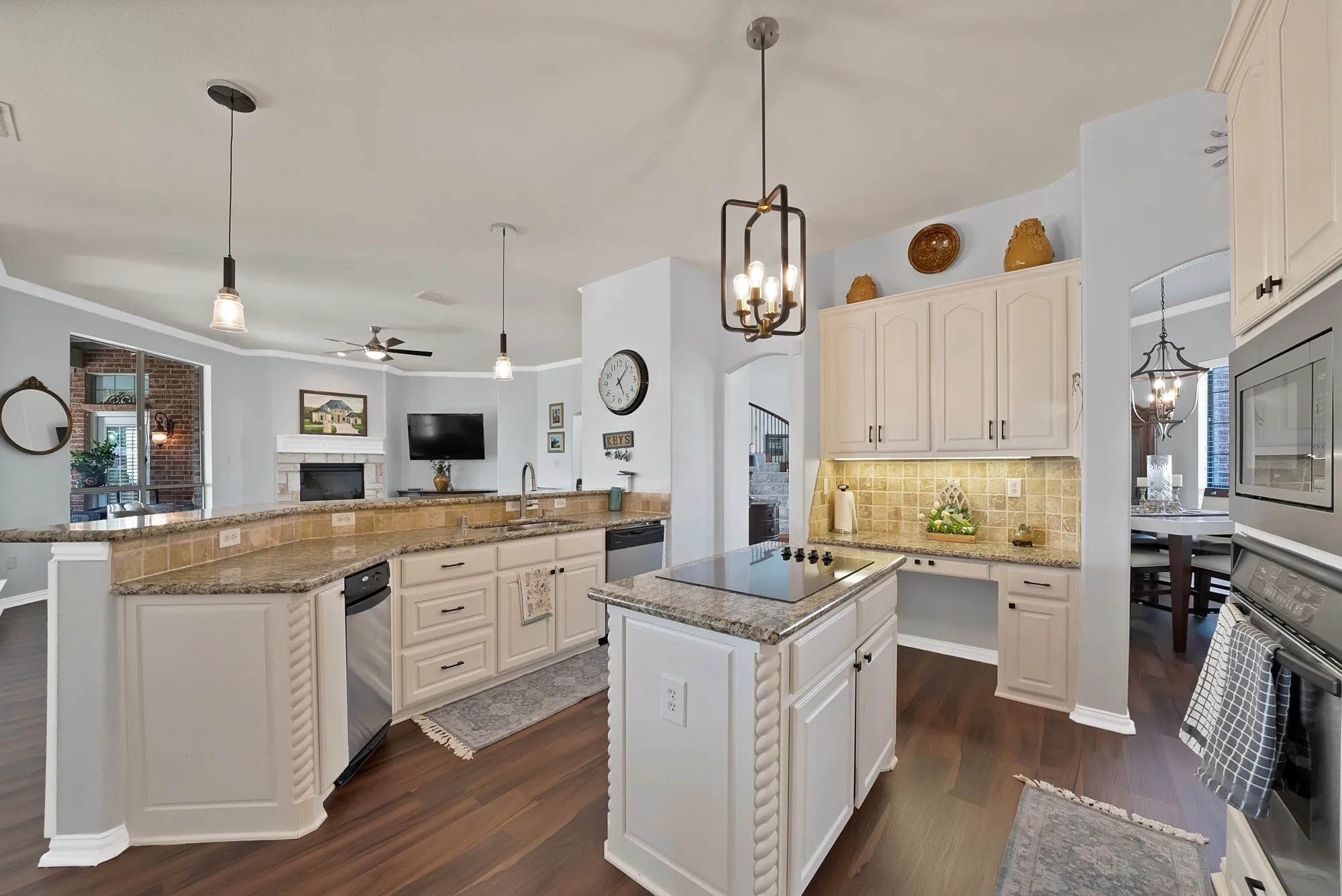 Kitchen — White raised-panel cabinetry, granite countertops with travertine tile backsplash, center island with cooktop, peninsula sink with open sightline to family room, and mixed pendant lighting including a brass lantern chandelier. Dark hardwood floors throughout.