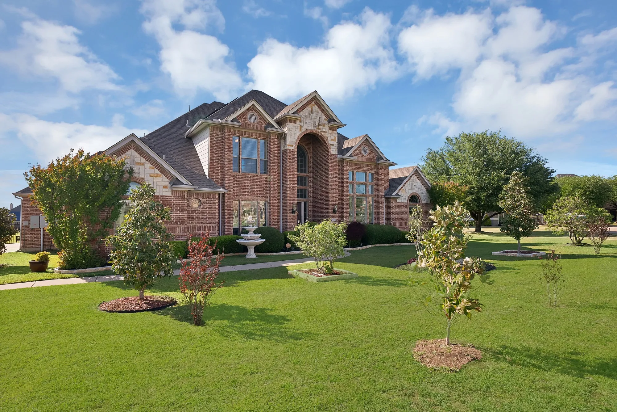 Front exterior, angle view — Brick and cut stone elevation with multi-gabled roofline, arched accent windows, and mature landscaping across an oversized front yard.