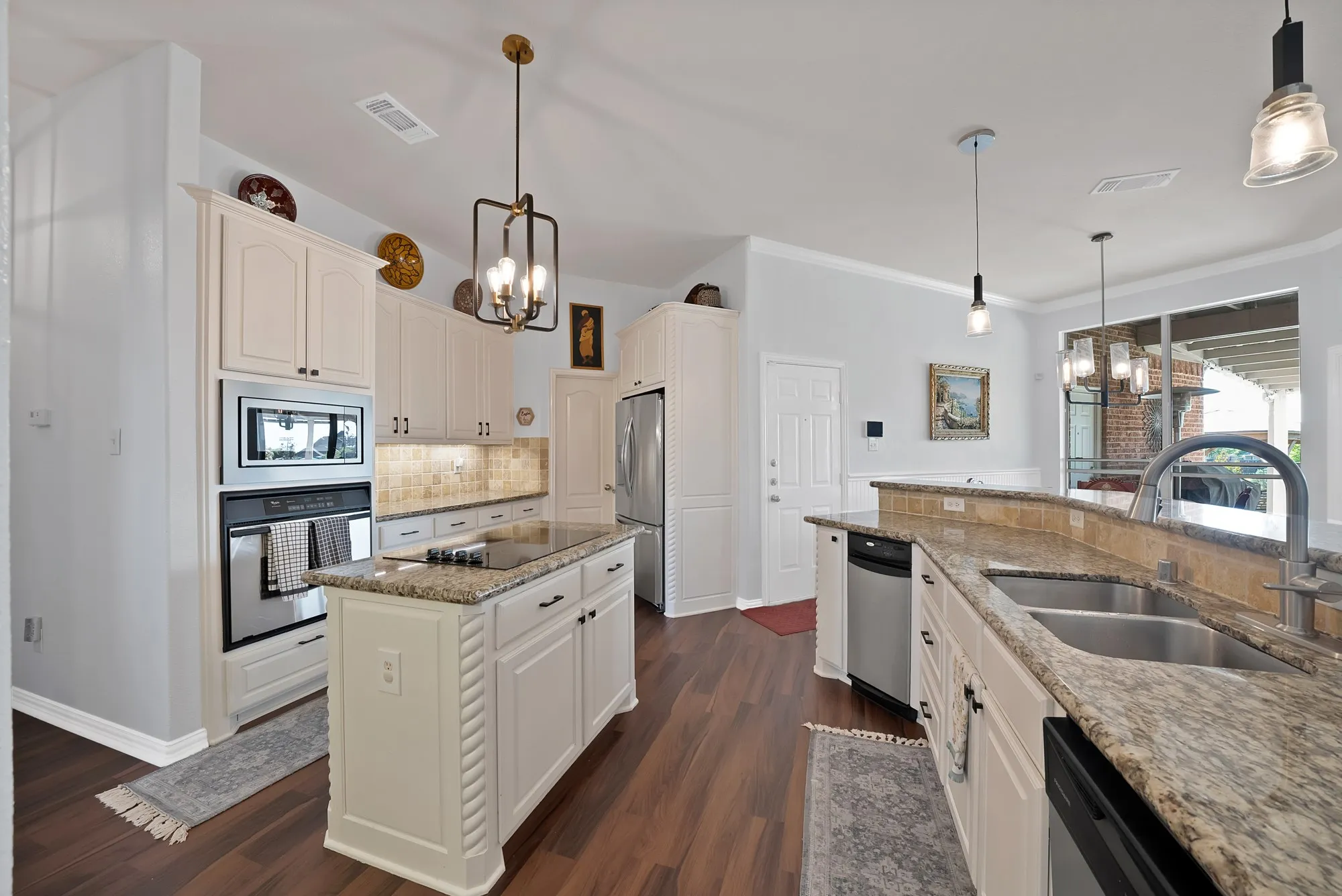 Kitchen, third angle — Full appliance wall with double wall oven, built-in microwave, stainless dishwasher, and undermount double sink. Island cooktop and granite perimeter countertops visible from this angle with direct sightline to covered back patio.