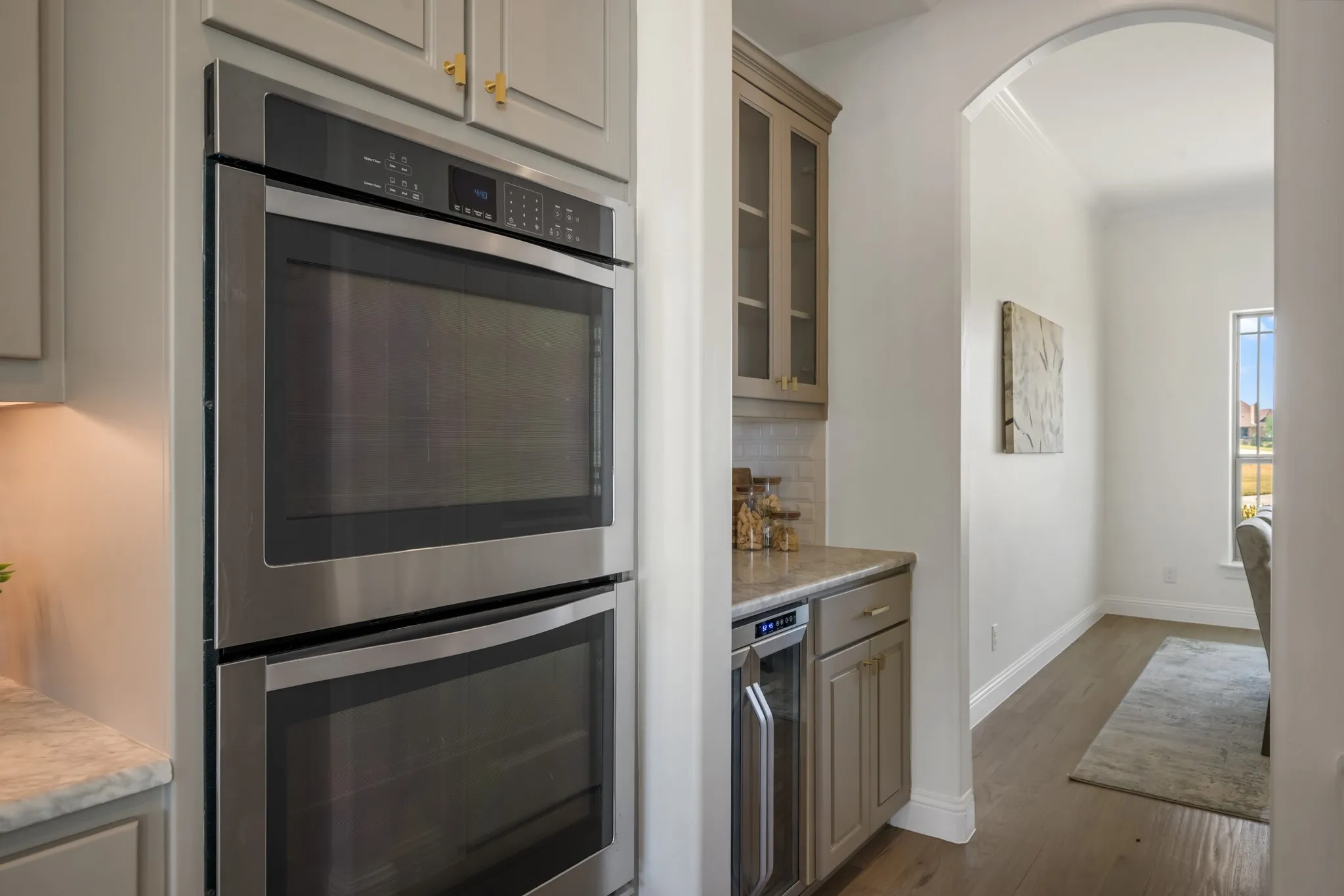 Built-in stainless steel double ovens, cabinetry with gold-tone hardware, a beverage cooler, light-toned countertops, and an arched entryway