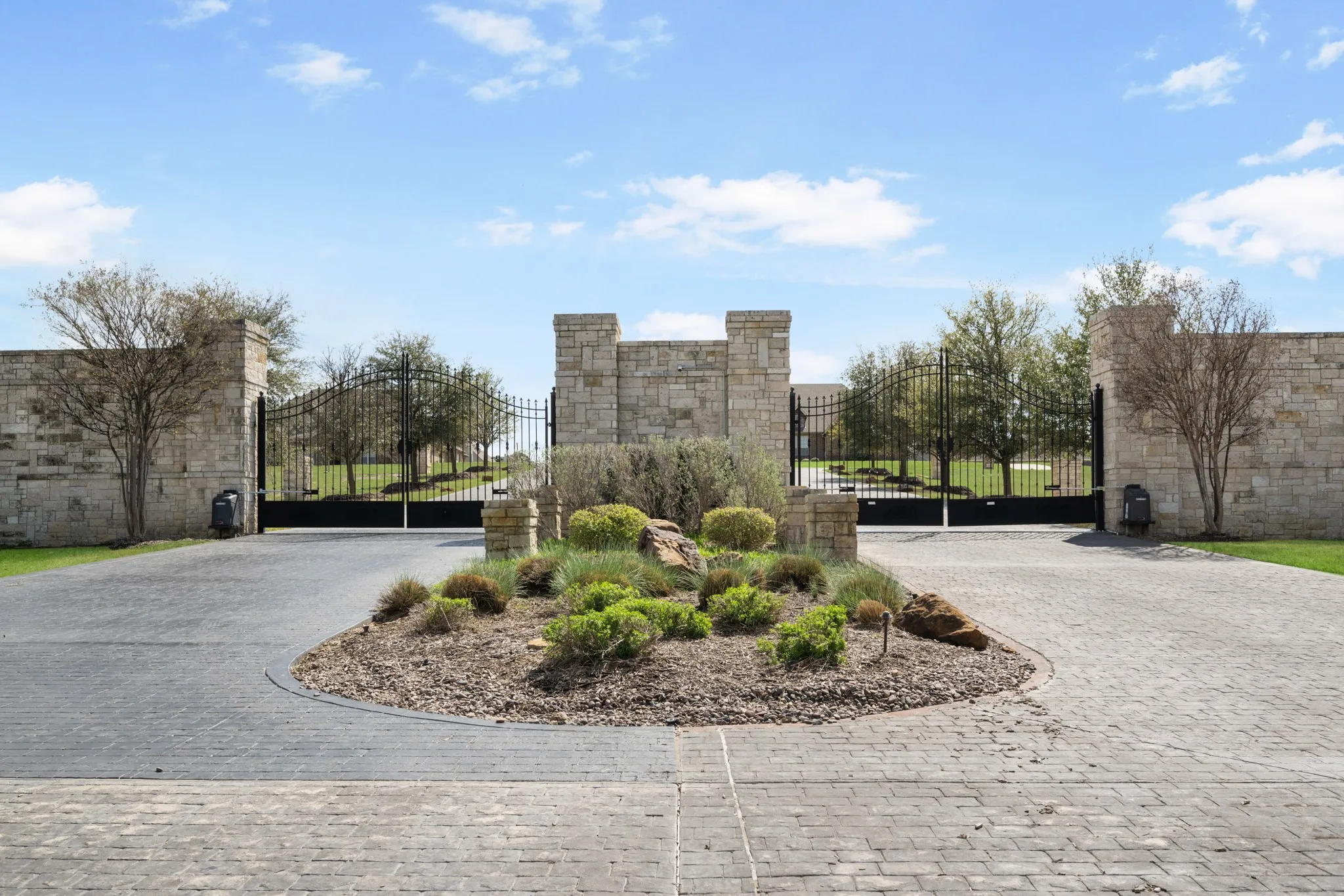 Stone entry columns with wrought iron gates, paver driveway, and a landscaped island
