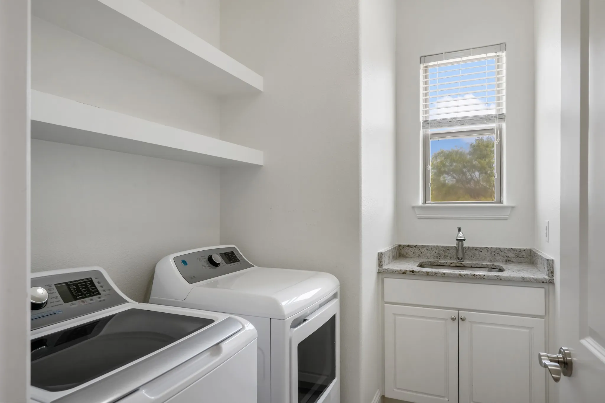 Laundry room featuring a single basin sink, granite-style countertop, white cabinetry, and two floating shelves