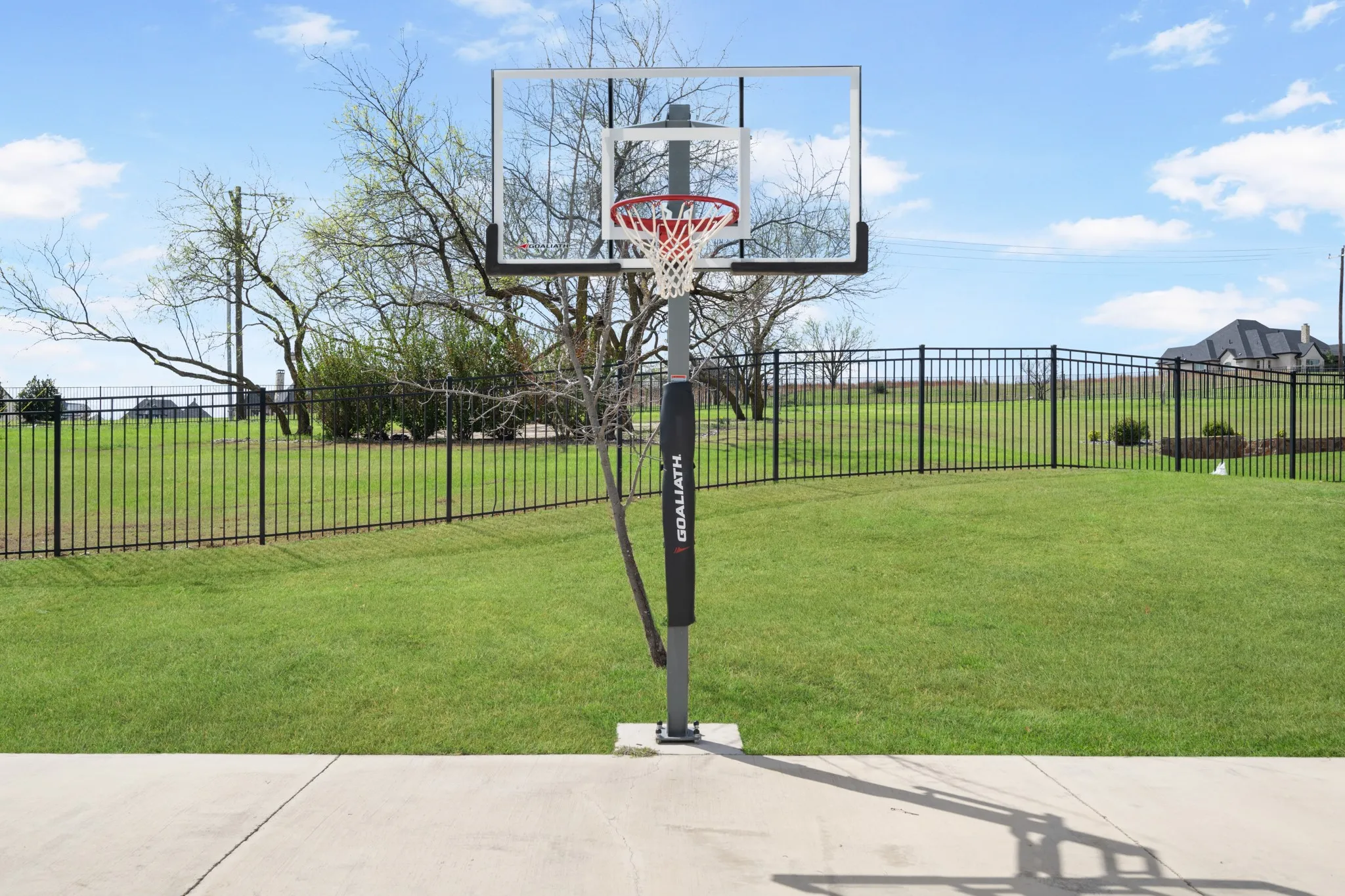 Expansive concrete patio with a freestanding basketball hoop, bordered by a substantial grassy yard and black metal fencing