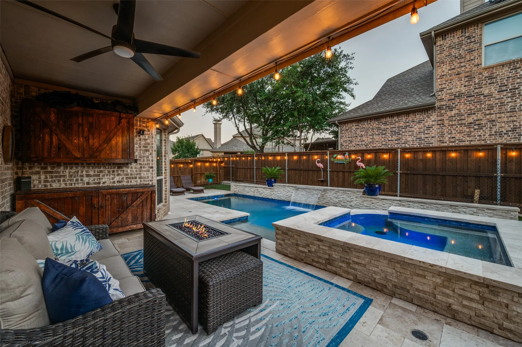 Resort-style backyard oasis featuring a spacious covered patio with a mounted TV, ceiling fan, and cozy lounge area overlooking the sparkling pool and raised spa.