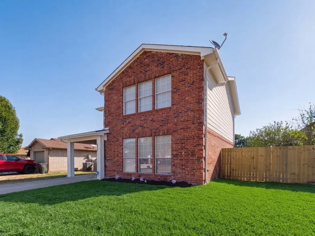 Brick exterior with white trim, covered carport, perimeter fencing, and a manicured lawn