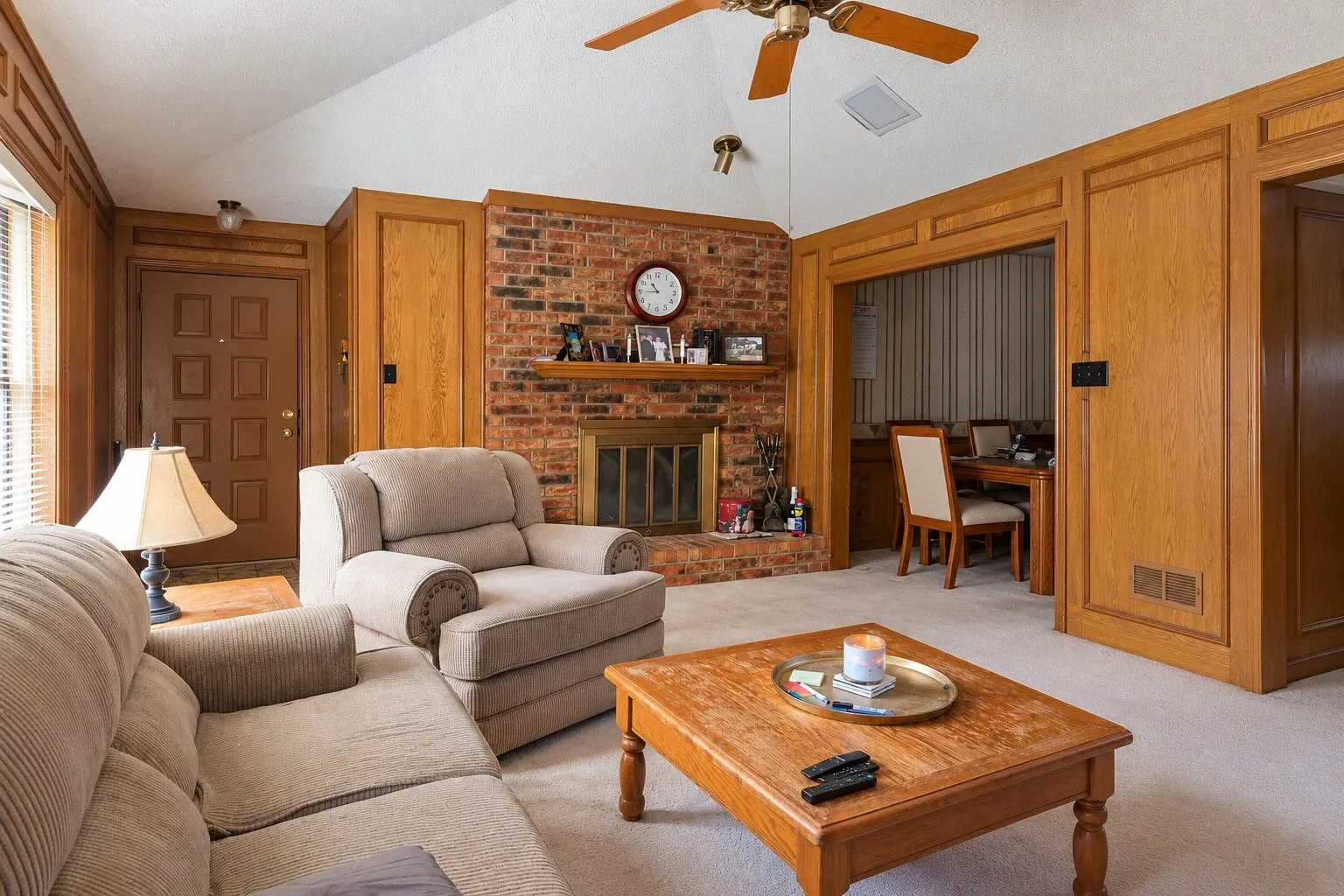 Vaulted ceiling with a ceiling fan, brick fireplace with a wood mantel, wood paneling, and a panel door entry