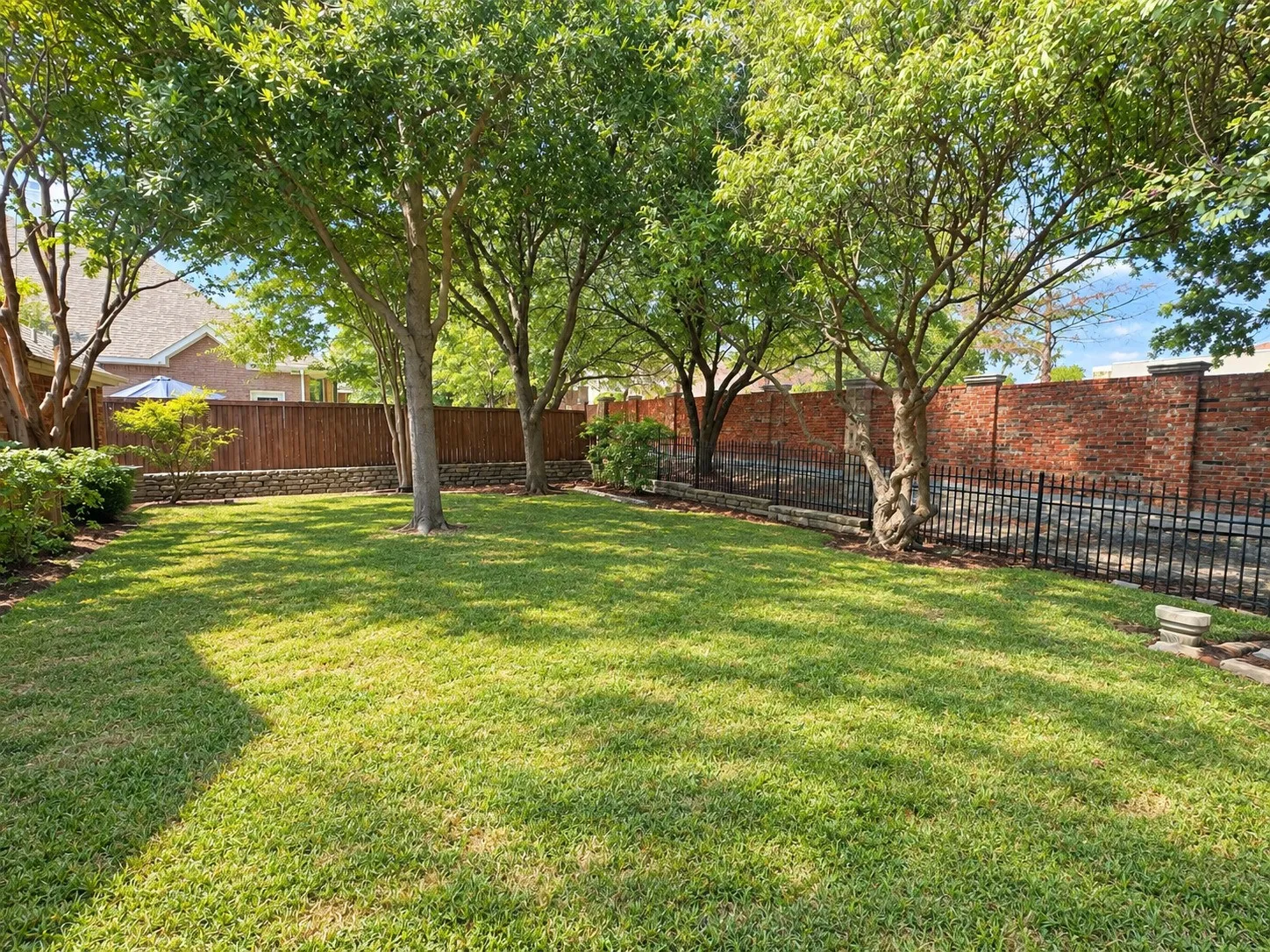 Expansive lawn area with mature trees, bordered by a dark wood fence, stone retaining wall, and brick wall
