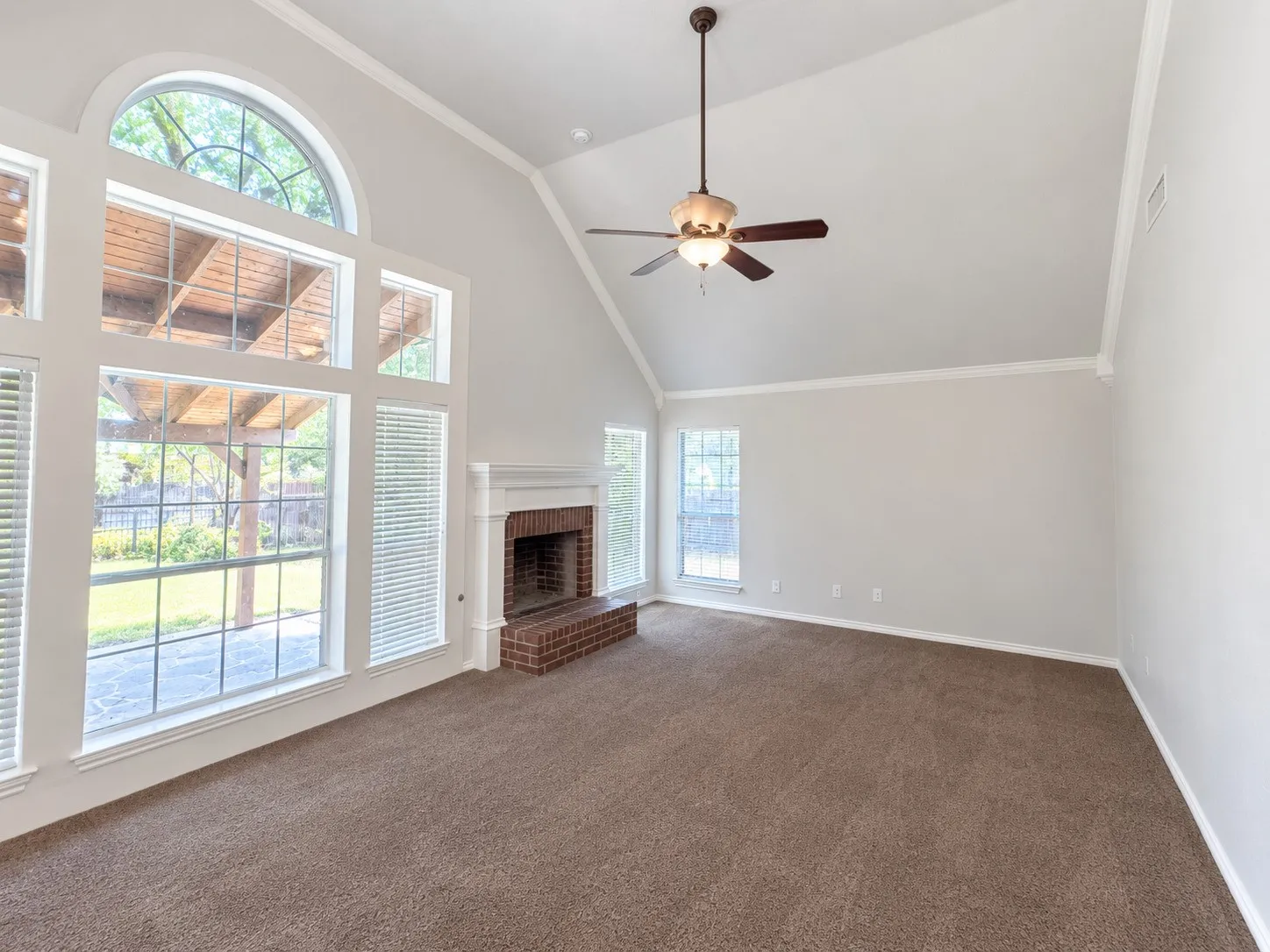 Vaulted ceiling, large arched window, brick fireplace with white mantel, carpet flooring, and a ceiling fan with integrated lighting