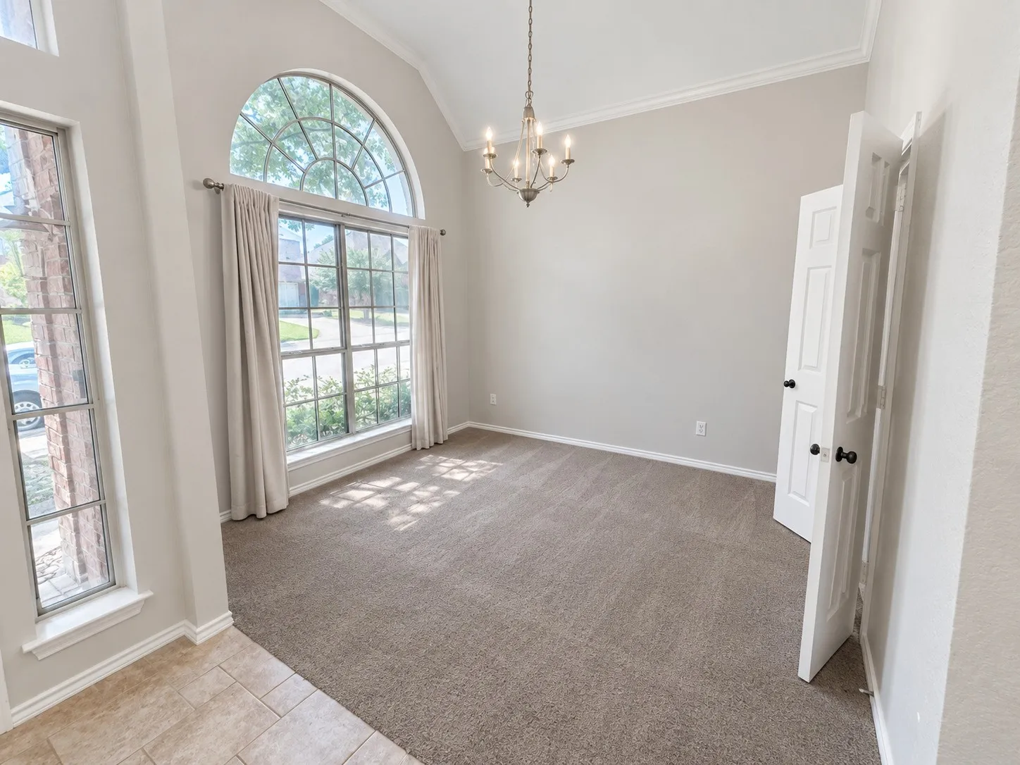 Vaulted ceiling with crown molding, arched window with grid pattern, chandelier lighting fixture, carpeted flooring, and a tile entry