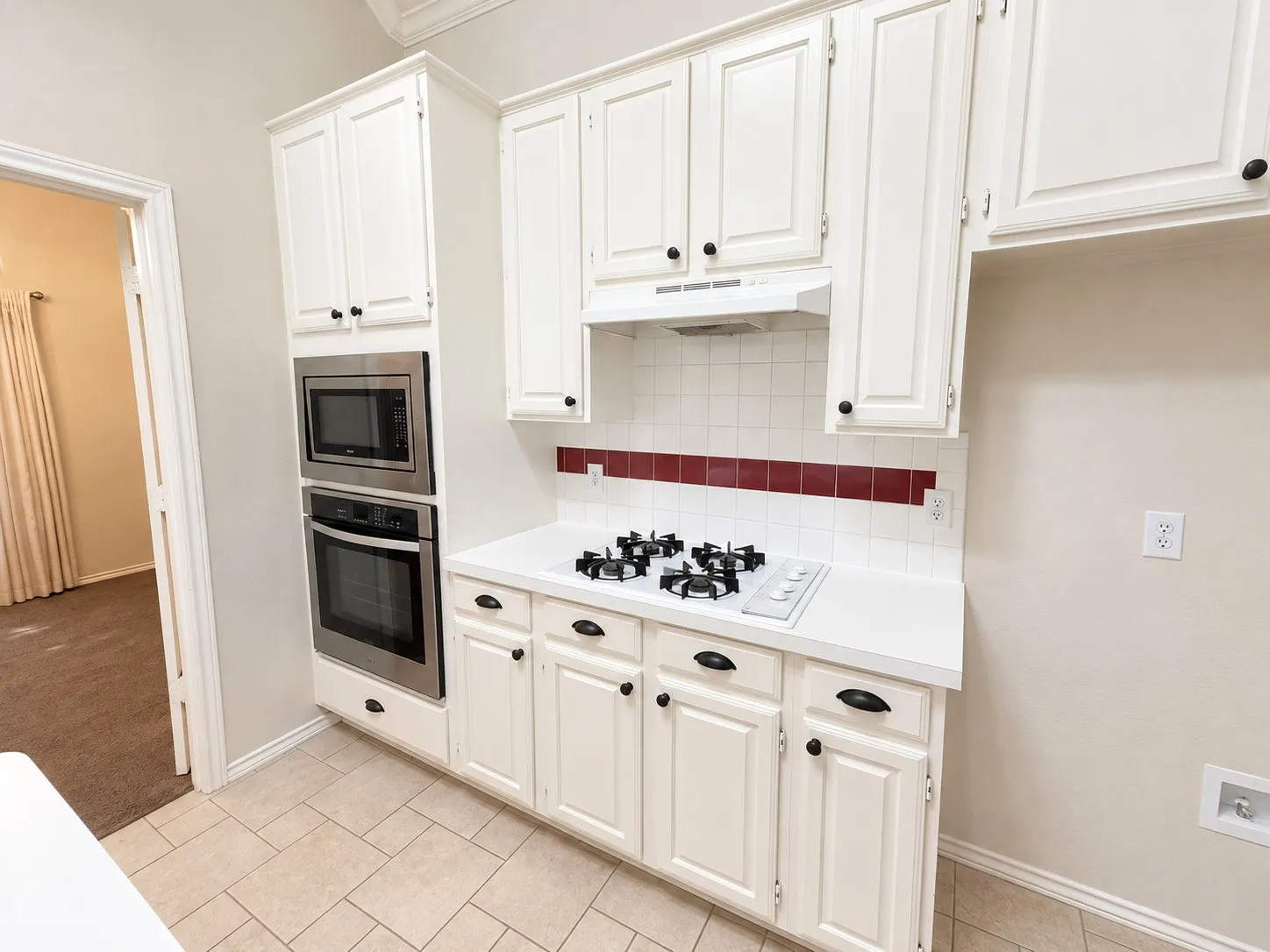 White cabinetry with black hardware, stainless steel built-in microwave and oven, gas cooktop, white tile backsplash with red accent, and neutral tile flooring