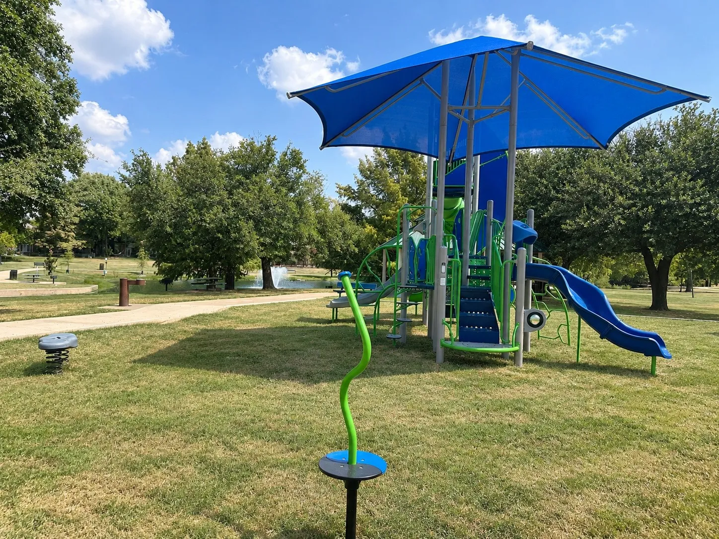 Expansive green space featuring a large playground with slides and a climbing structure, shaded by a blue​​‌​​​​‌​​‌‌​‌‌​​​‌‌​‌​‌​‌​​​‌​​ canopy