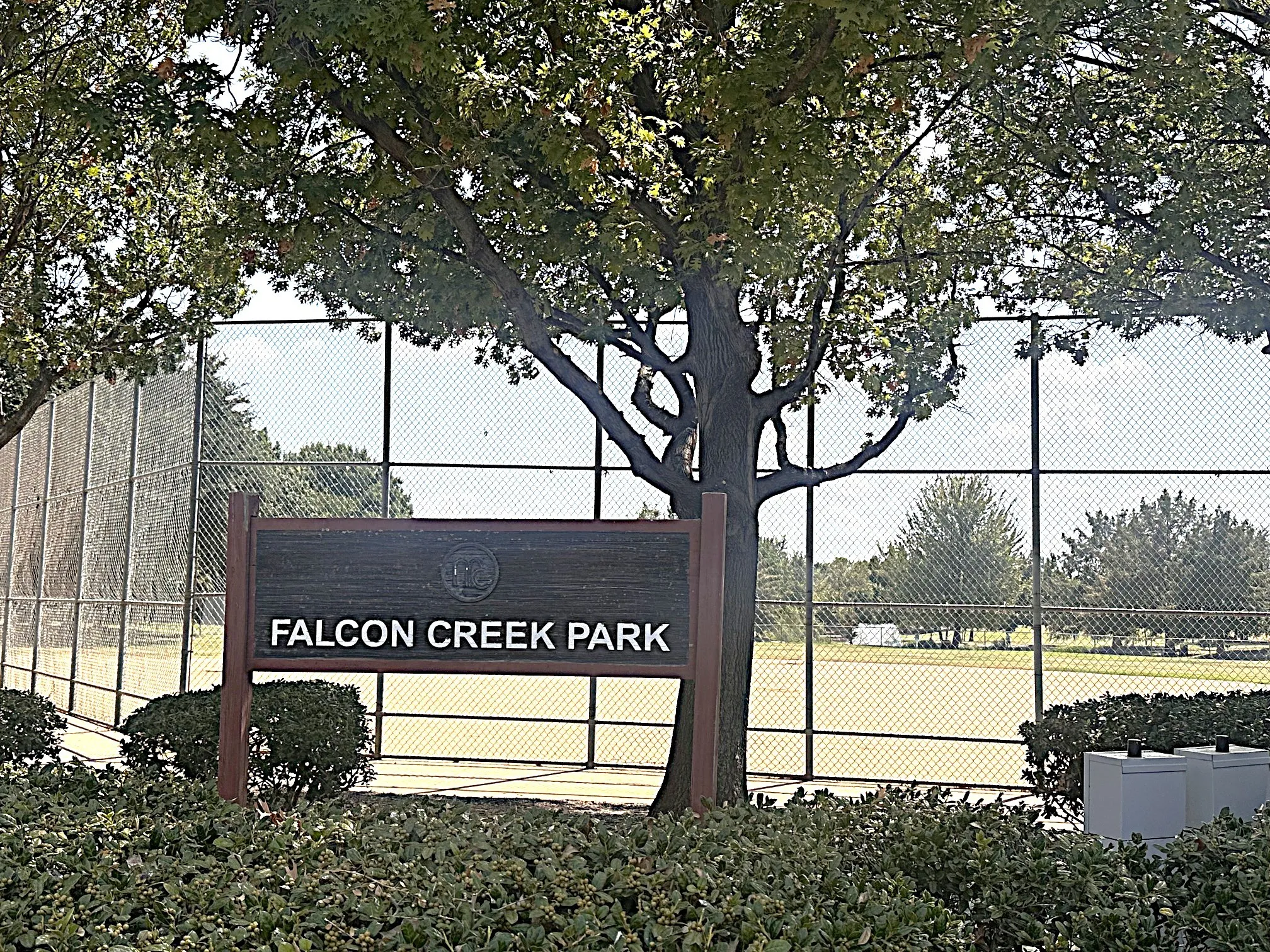 Falcon Creek Park signage, surrounded by mature trees and manicured shrubbery, with a fenced sports court visible in the background