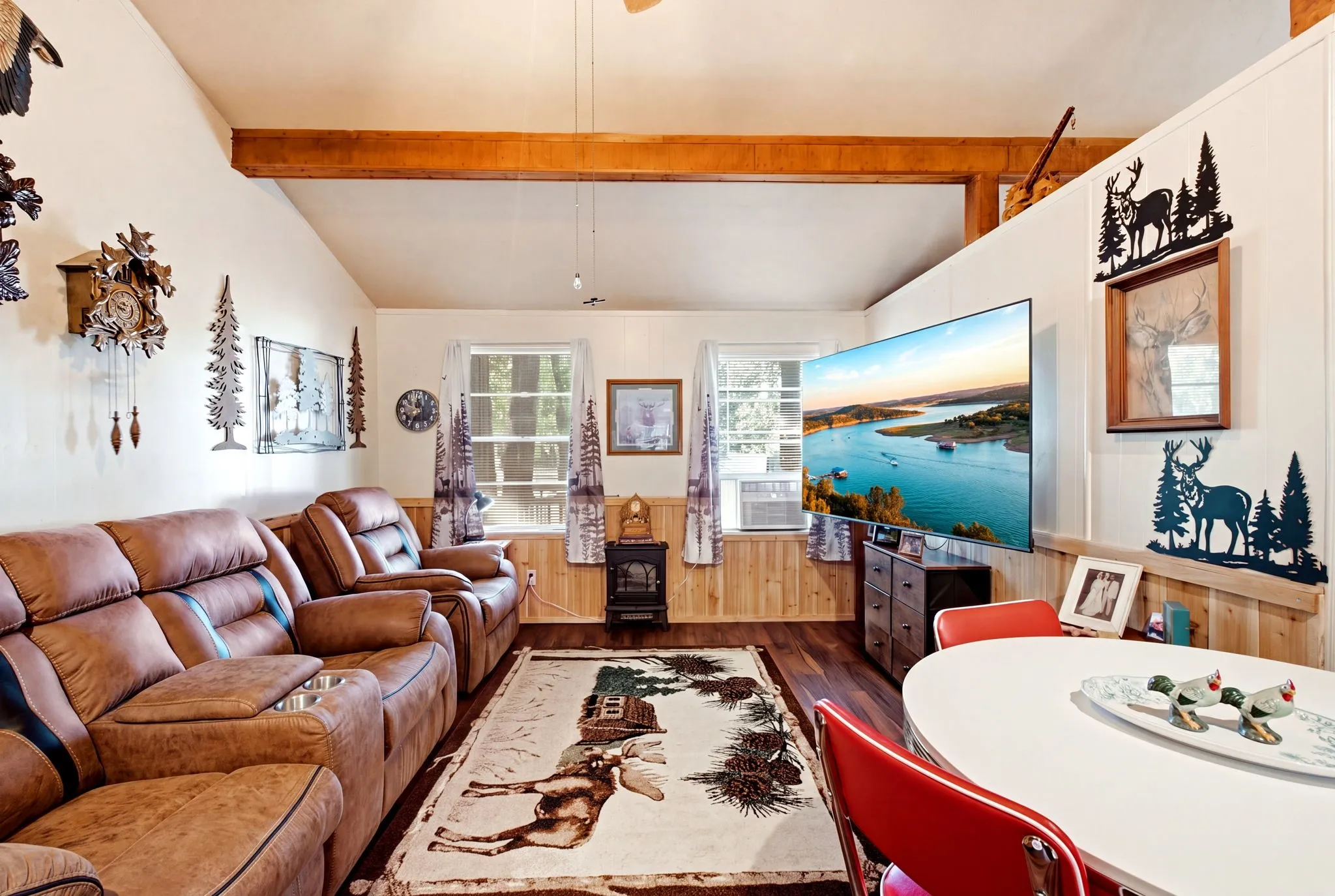 Vaulted ceiling with exposed wood beams, wood-finish flooring, two large windows, wood paneling, and a compact built-in fireplace