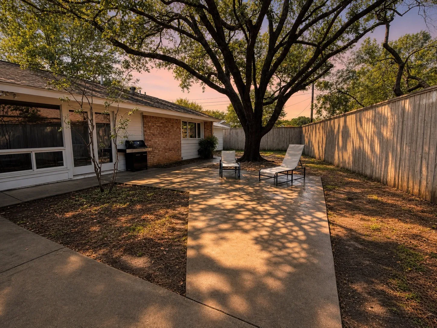 Private backyard featuring an expansive concrete patio, mature trees providing natural shade, a wood privacy fence, and brick exterior accents on the home