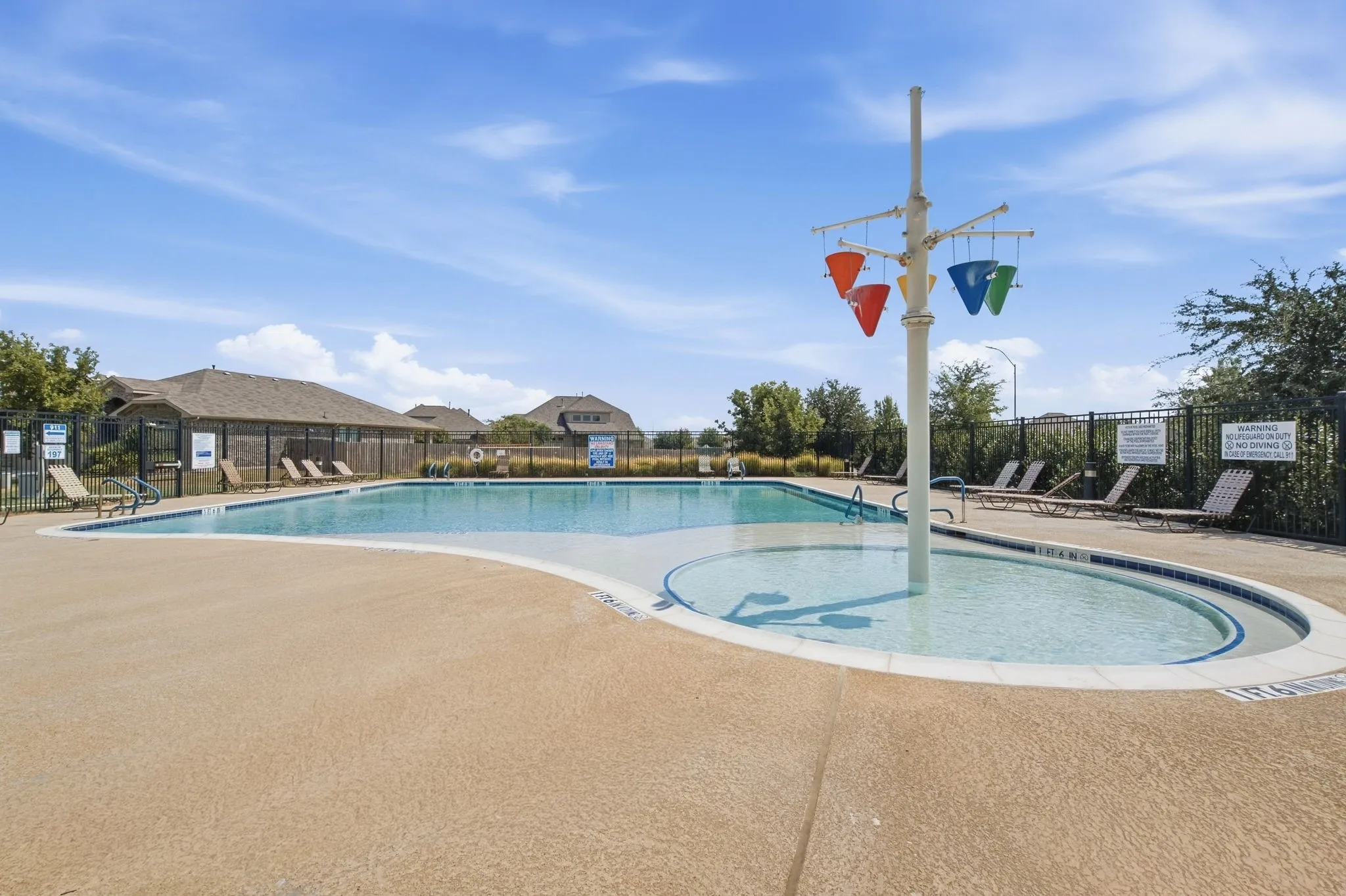 Community swimming pool featuring a shallow water play area with a water feature, surrounding concrete decking, and perimeter fencing