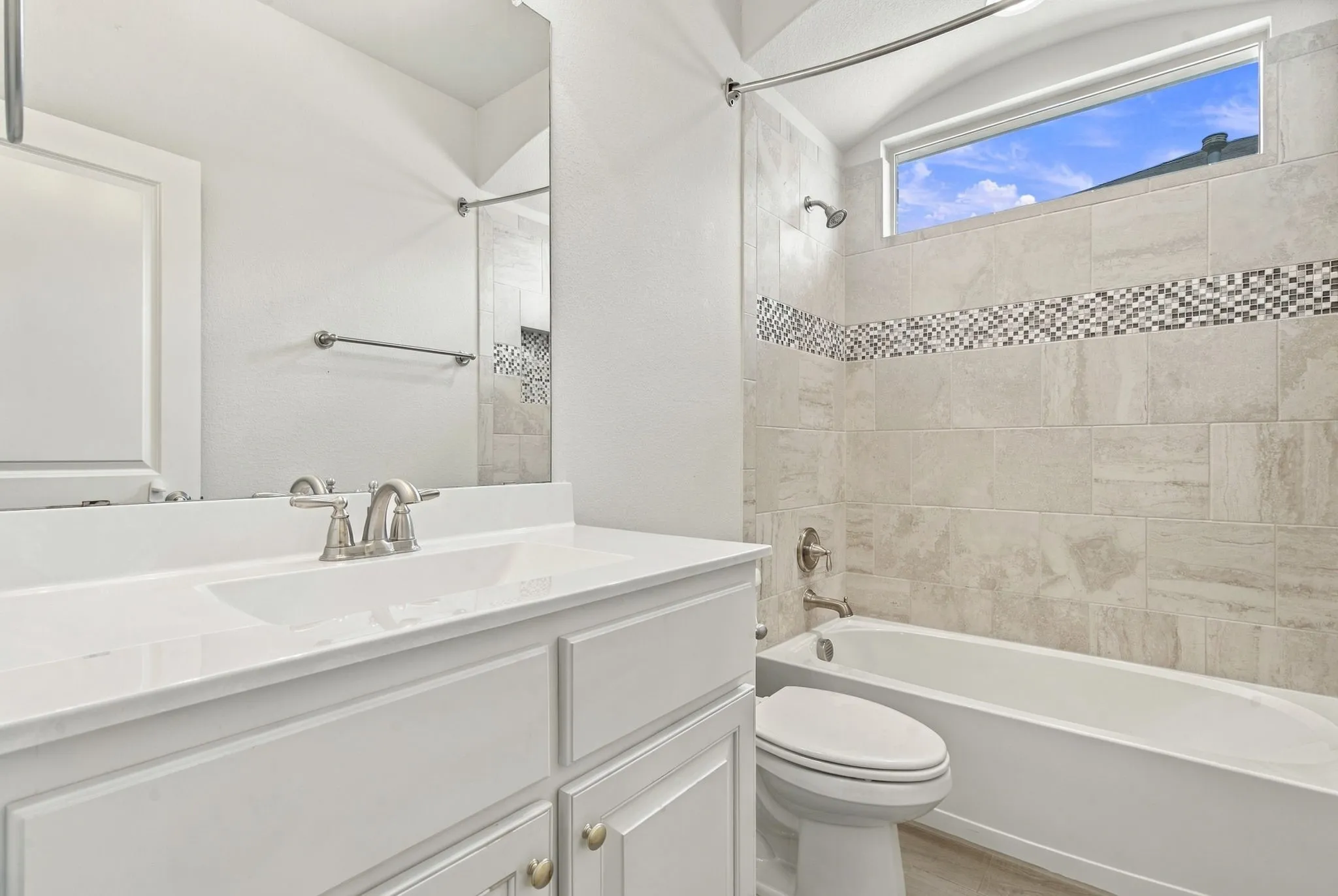 White vanity with integrated sink, brushed nickel faucet, full-width mirror, subway tiled bath/shower combination with mosaic accent, and arched window