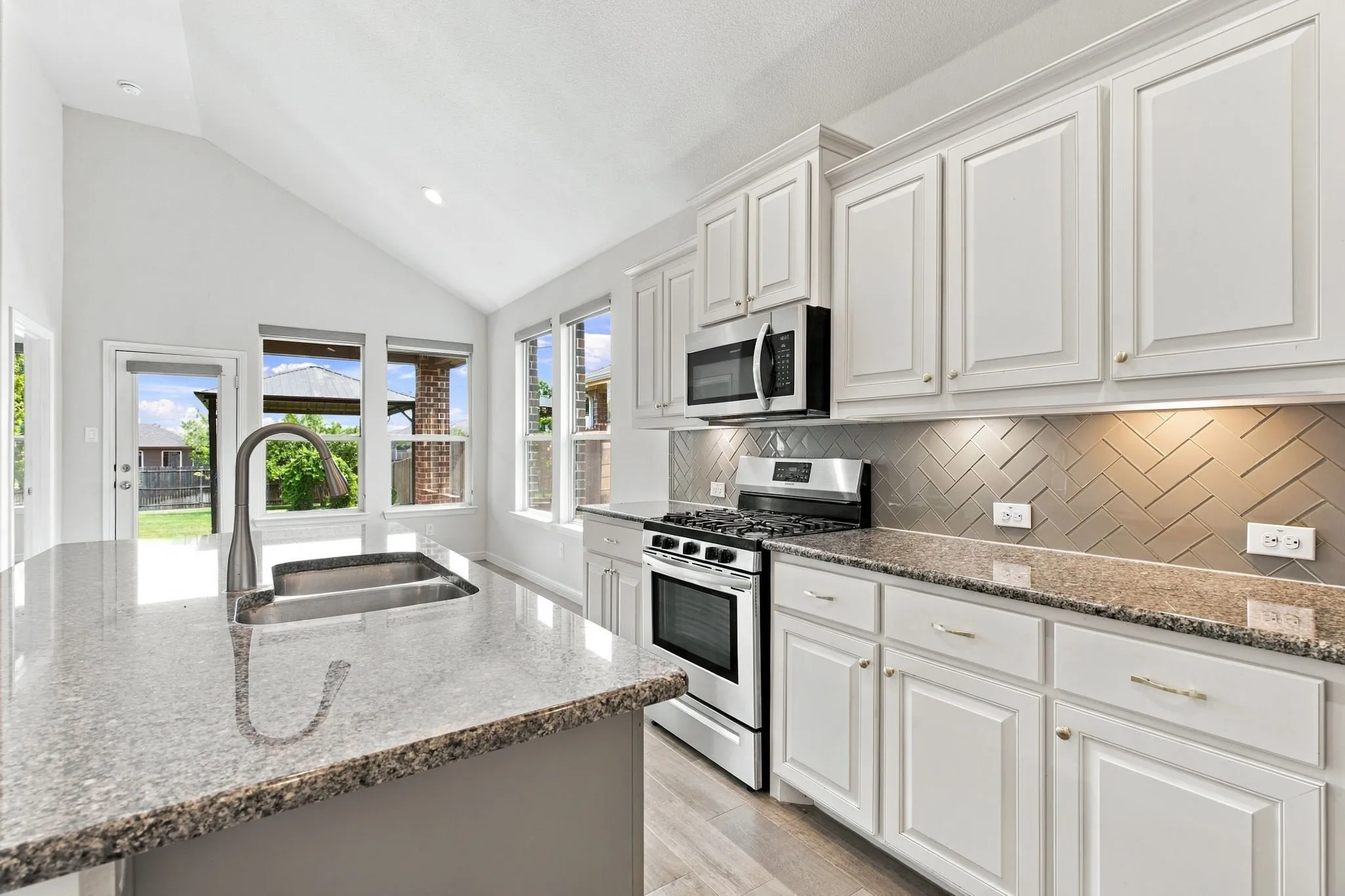 Vaulted ceiling kitchen featuring white cabinetry, stone countertops, stainless steel appliances, and a herringbone tile backsplash