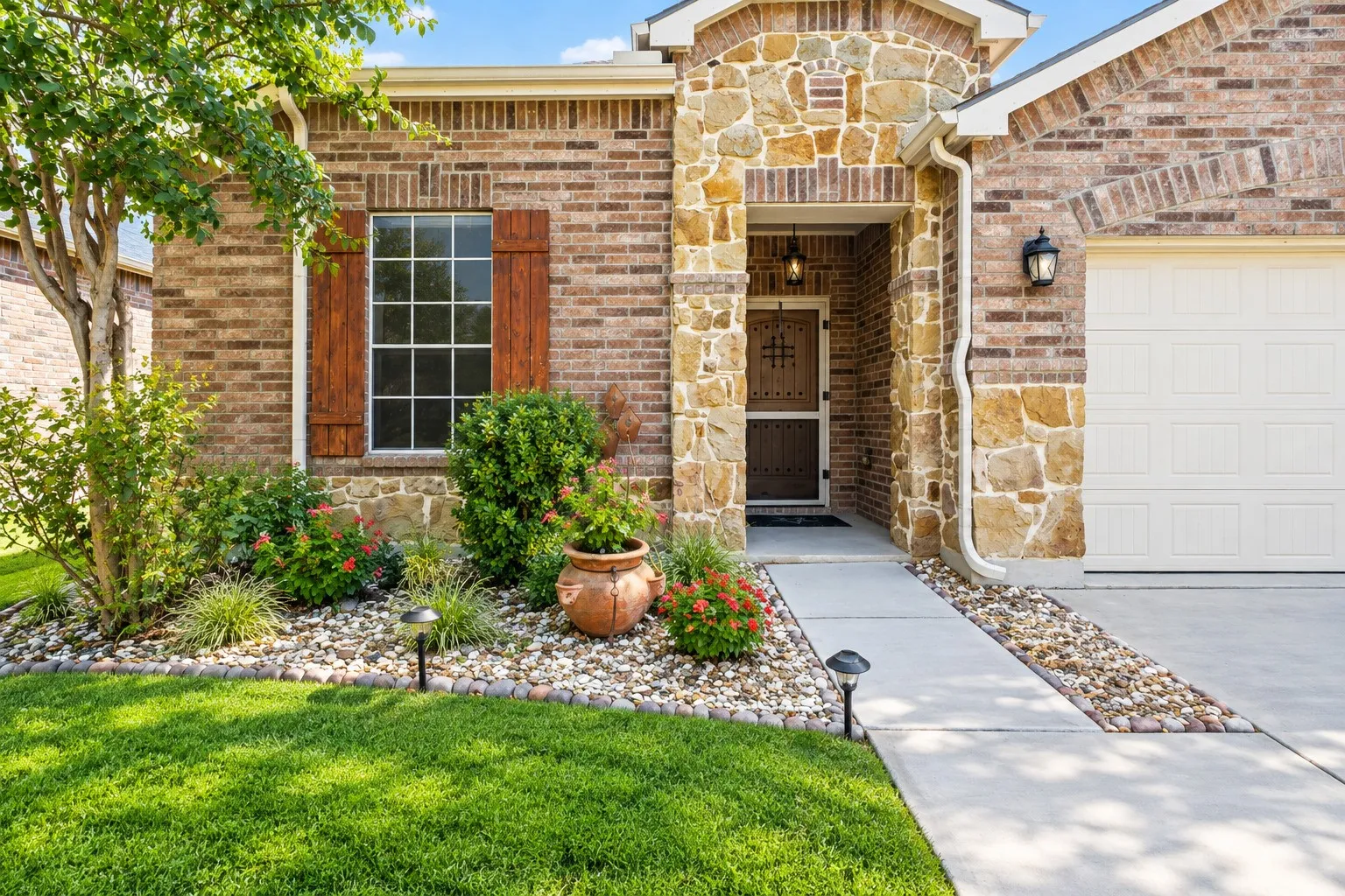 Brick and stone exterior with decorative wood shutters, lush landscaping, concrete walkway, and attached​​‌​​​​‌​​‌‌​‌‌​​​‌‌​‌​‌​‌​​​‌​​ garage