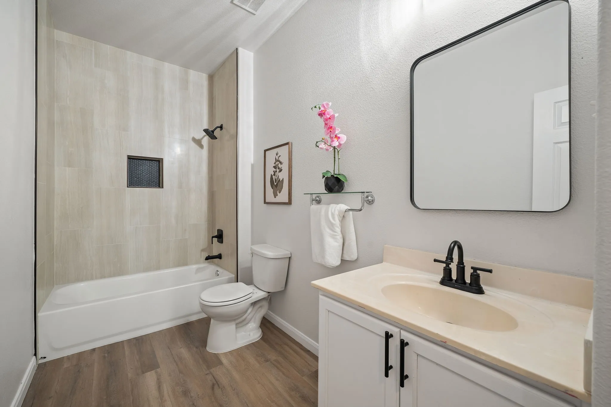 Bathroom featuring a shower-tub combination with light-toned tiled surround, wood-finish flooring, a vanity with a light-toned countertop, and a contemporary wall mirror