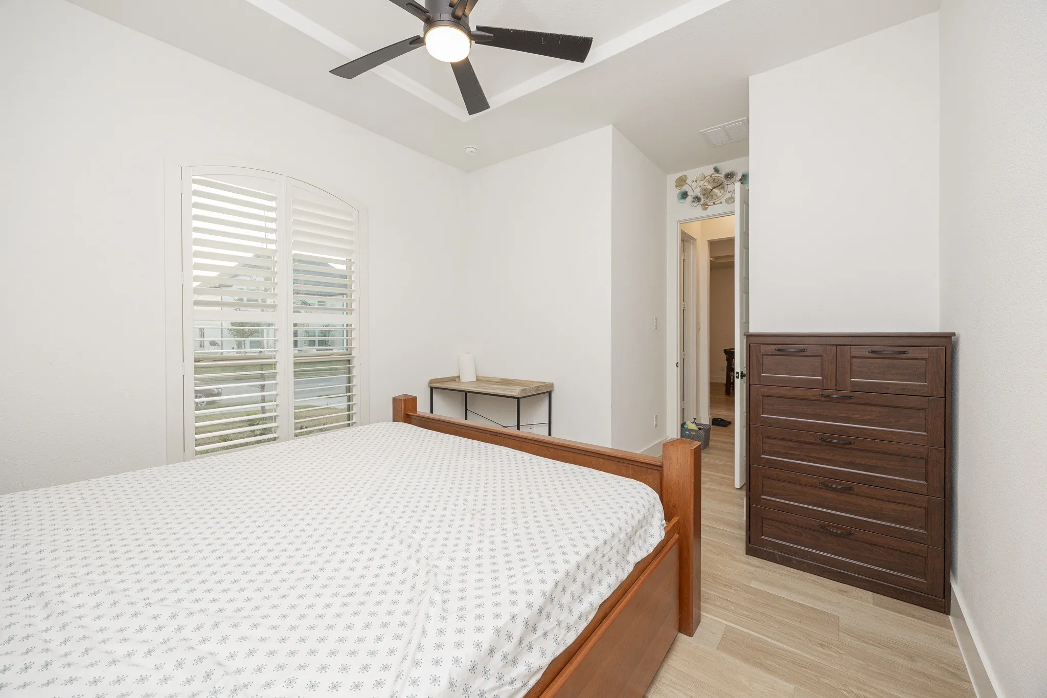 Arched window with plantation shutters, ceiling fan with integrated lighting, tray ceiling, wood-finish flooring