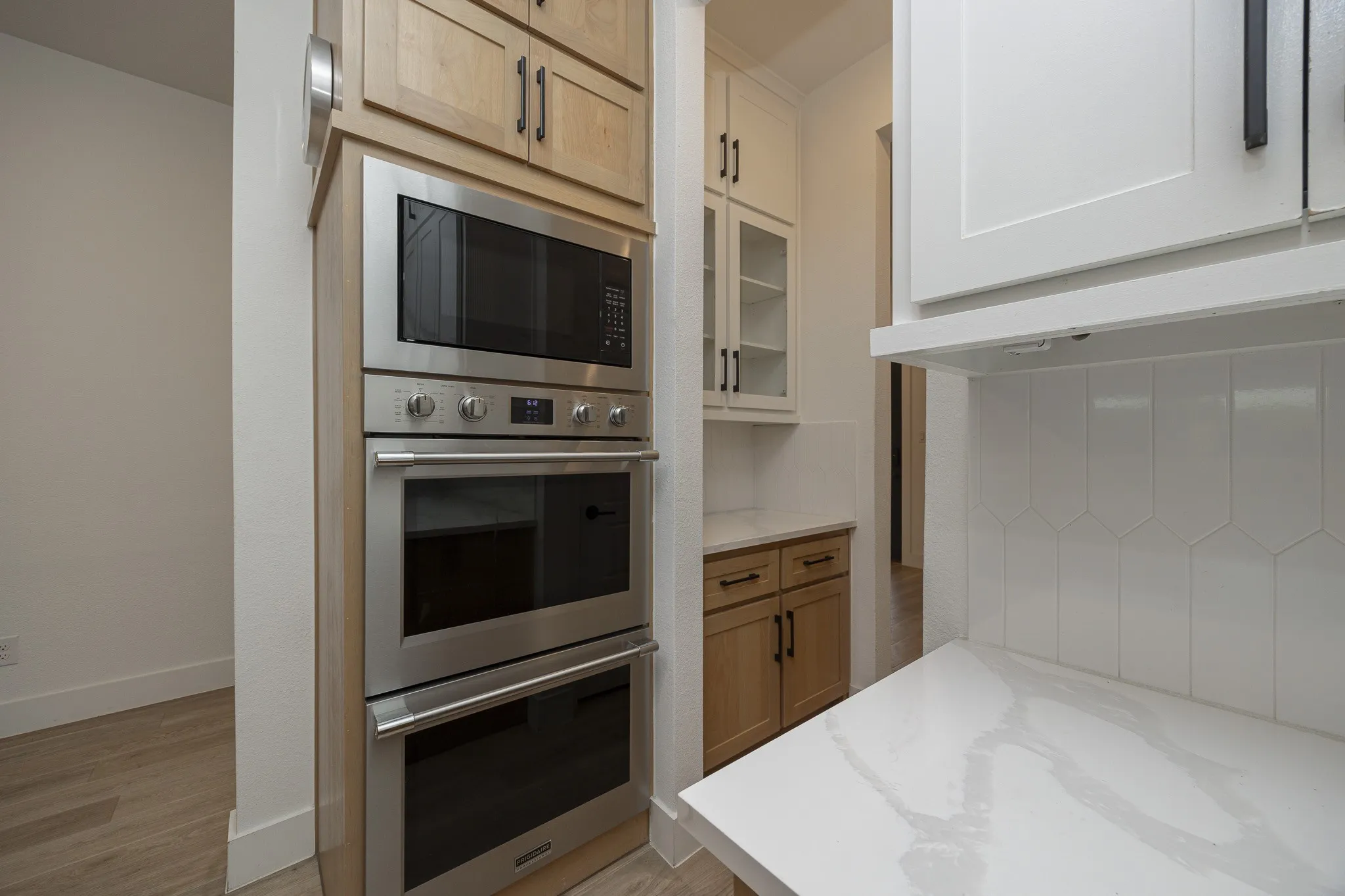 Built-in stainless steel oven and microwave combination, light wood-finish cabinetry, white countertop, and white backsplash with a geometric tile pattern